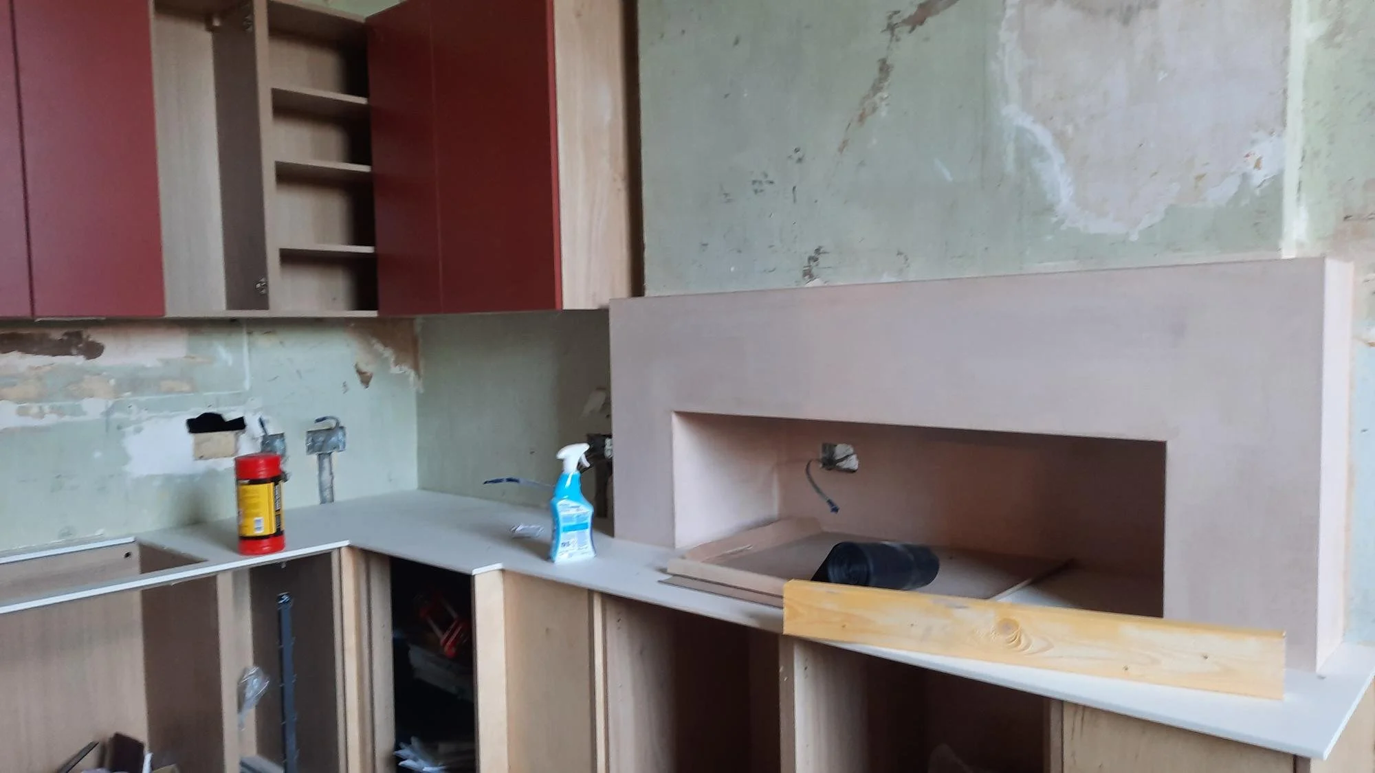 Kitchen under renovation with unfinished cabinetry and exposed drywall, a can of red primer, a spray bottle, and construction tools on the countertop.