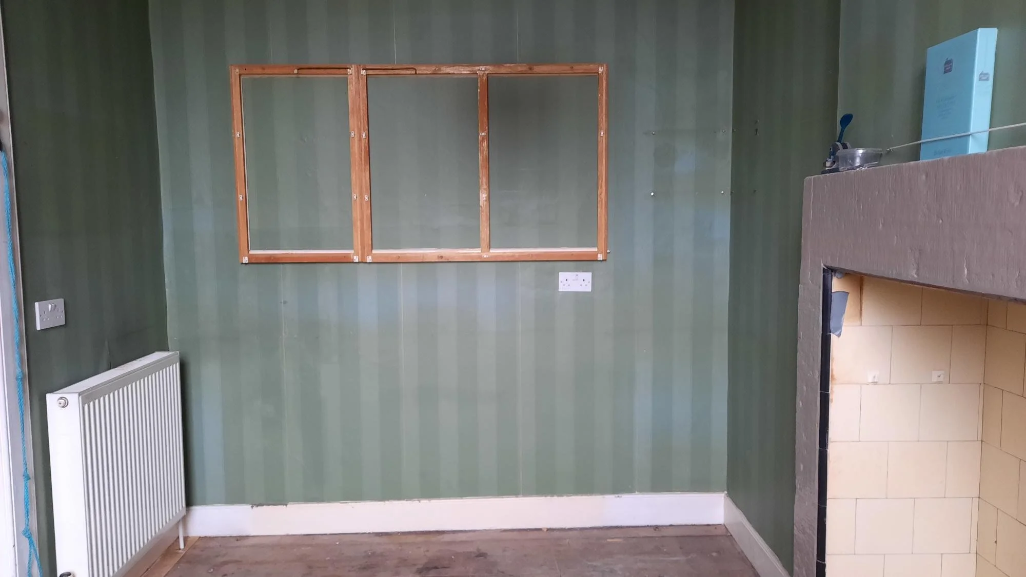 Interior of a room with green striped wallpaper, a radiator on the left wall, an unfinished wooden wall frame in the center, a double power socket beneath it, and part of a tiled fireplace or counter with a blue box and some tools on top on the right
