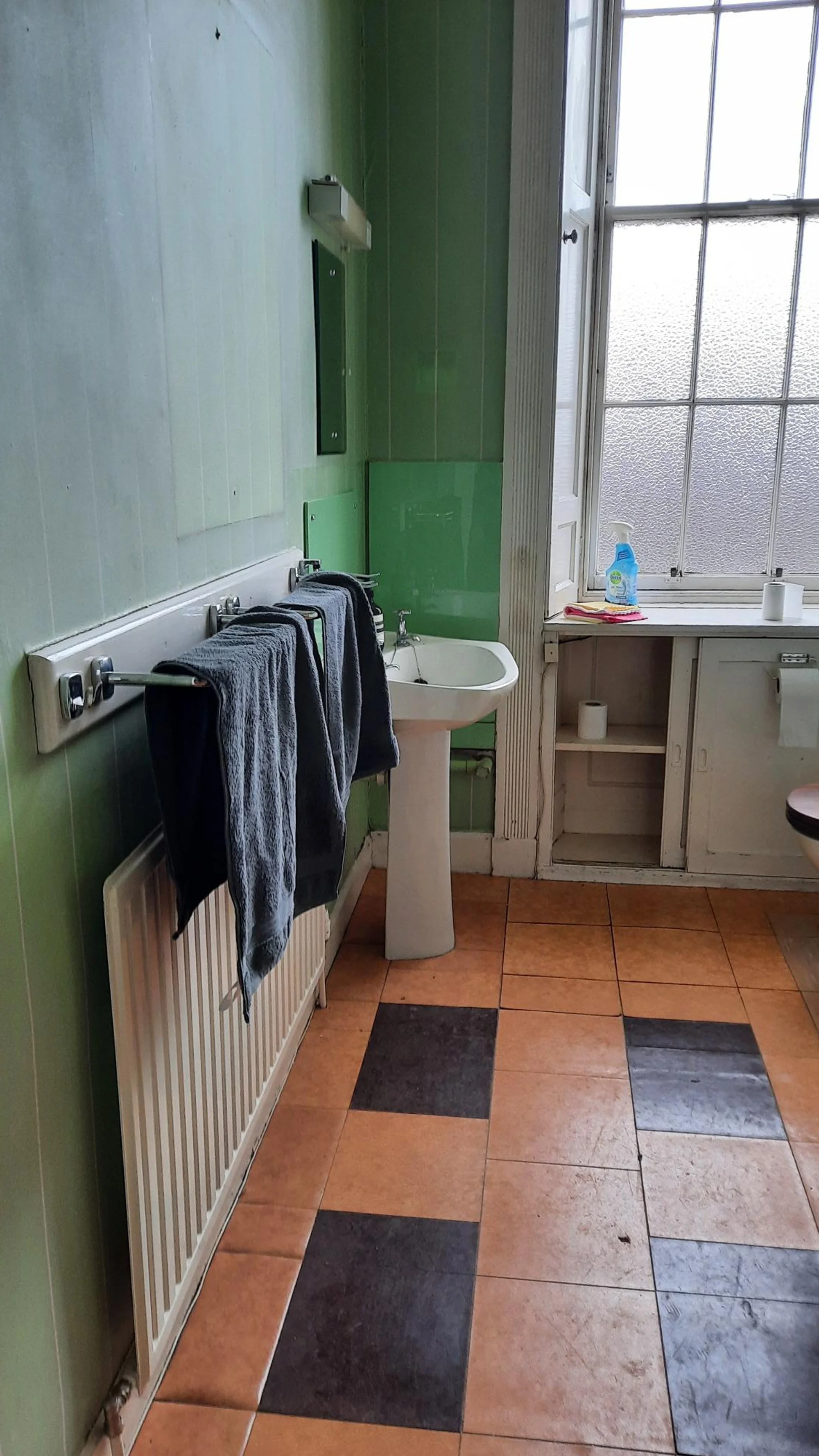 A bathroom with green tiled walls, an open window with frosted glass, a white pedestal sink, a black toilet seat, a towel rack with dark towels, and a small shelf with cleaning spray and toilet paper.