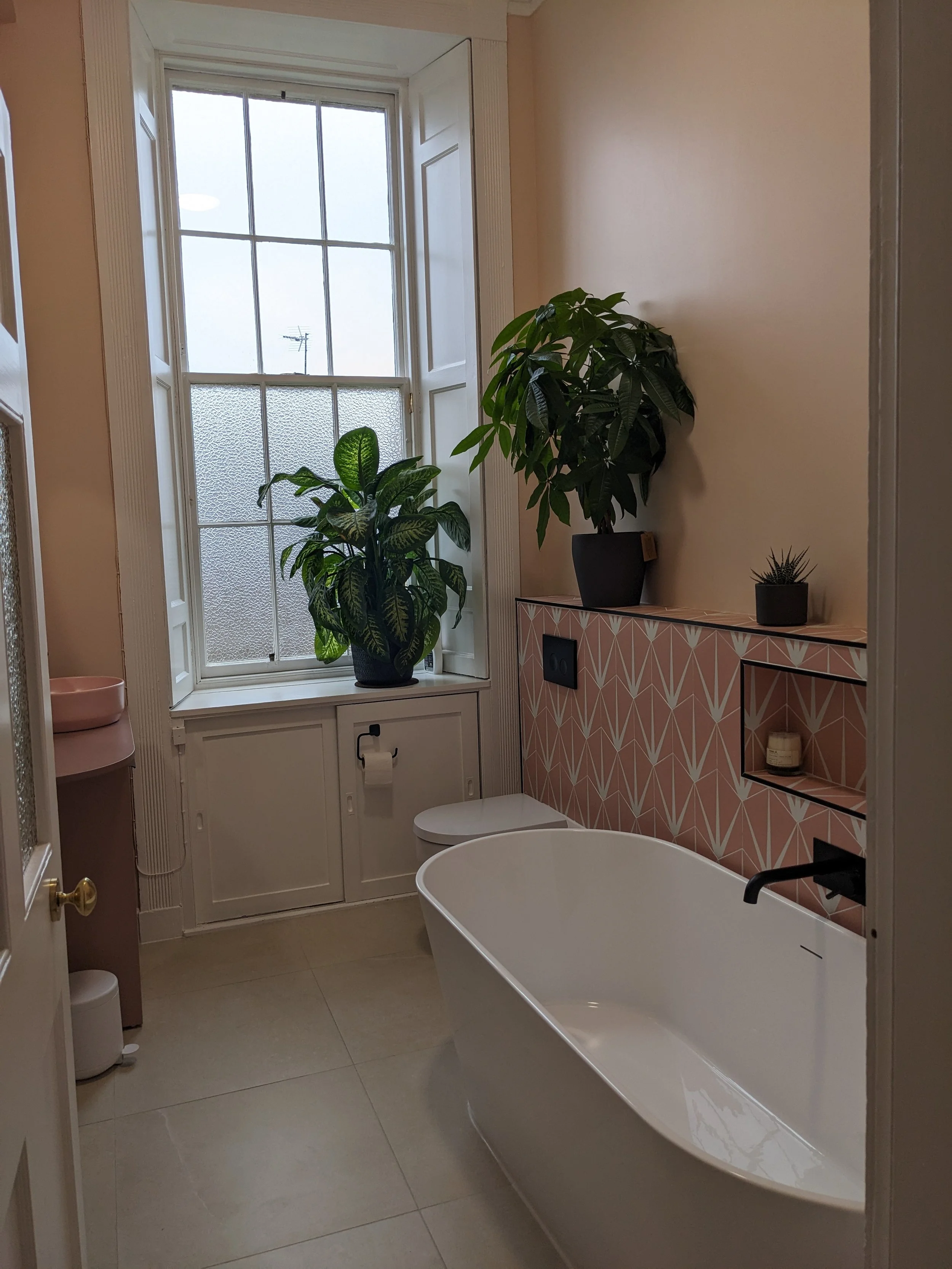 Modern bathroom with a freestanding bathtub, two large potted plants on a pink geometric-patterned wall, a window with frosted and clear glass panes, and small decorative items on the wall shelf.