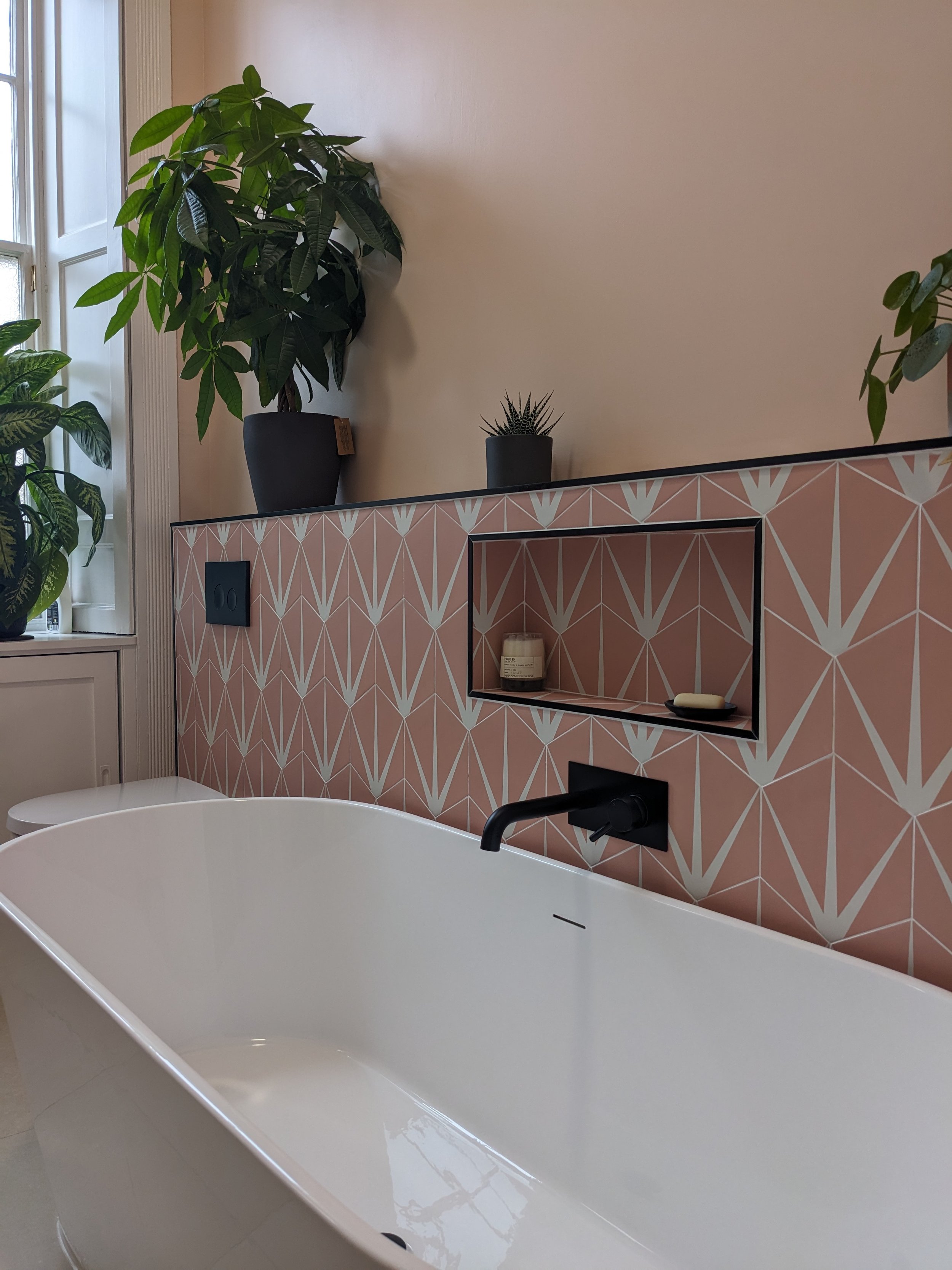 Modern bathroom with a white bathtub, black faucet, pink geometric patterned wall, and potted plants on the shelf.