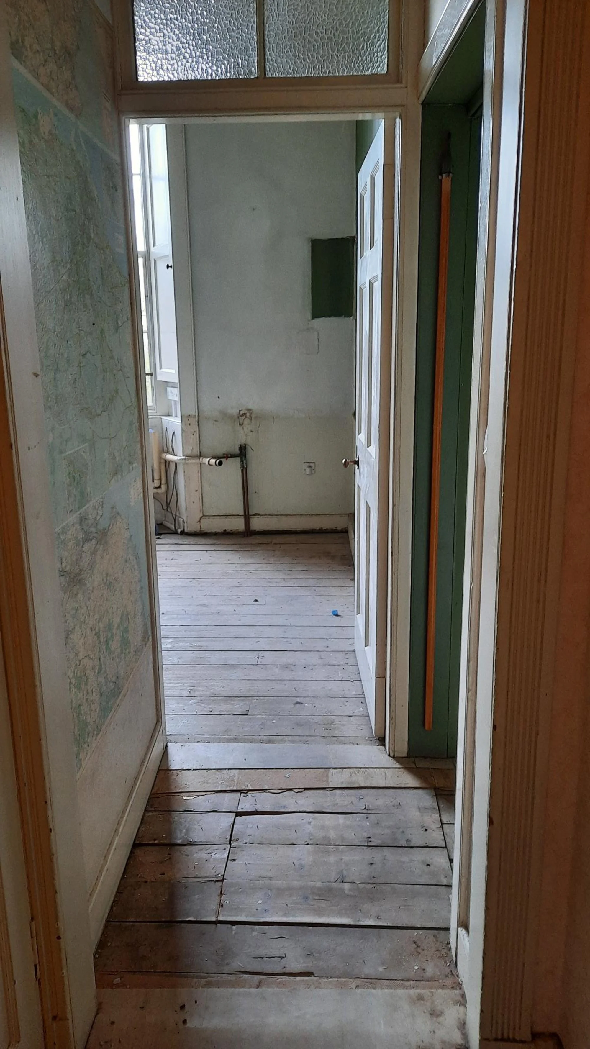 An empty, old hallway leading into a small room with a window and exposed pipes on the wall.