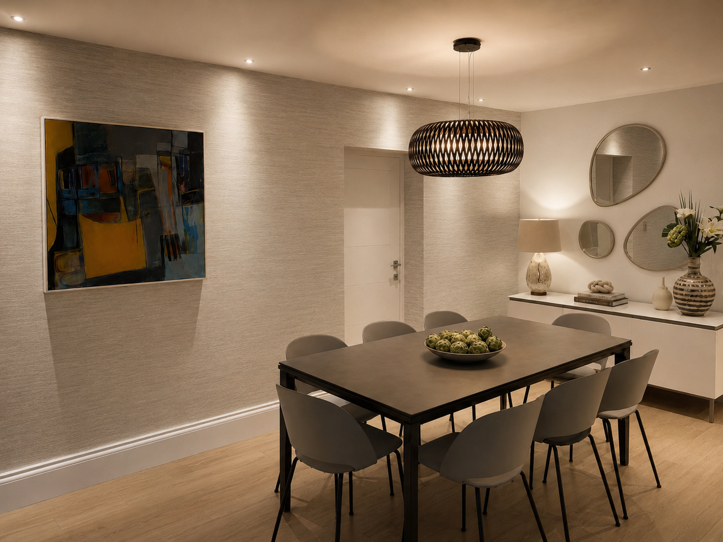 A modern dining room with a dark wood table surrounded by six gray chairs. A bowl of green decorative balls is on the table. On the back wall, there are three mirrors, a lamp, and decorative items on a white sideboard. A large abstract painting hangs