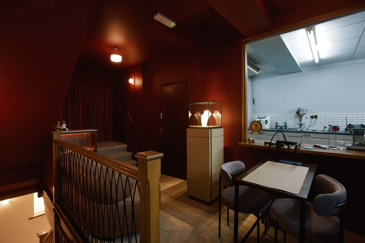A cozy interior space with a staircase, wooden wall panels, and a small dining table with four chairs. There is a glass display case with decorative lamps, and a kitchen area visible through a large open window with white subway tile backsplash and various appliances.