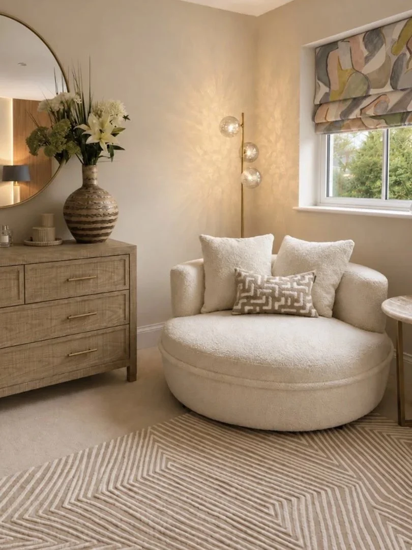 A cozy living room corner with a round cream sofa, decorative pillows, a wooden dresser with a large vase of white flowers and greenery, a modern floor lamp with three glass globes, a window with a patterned Roman shade, a mirror on the wall, and a g