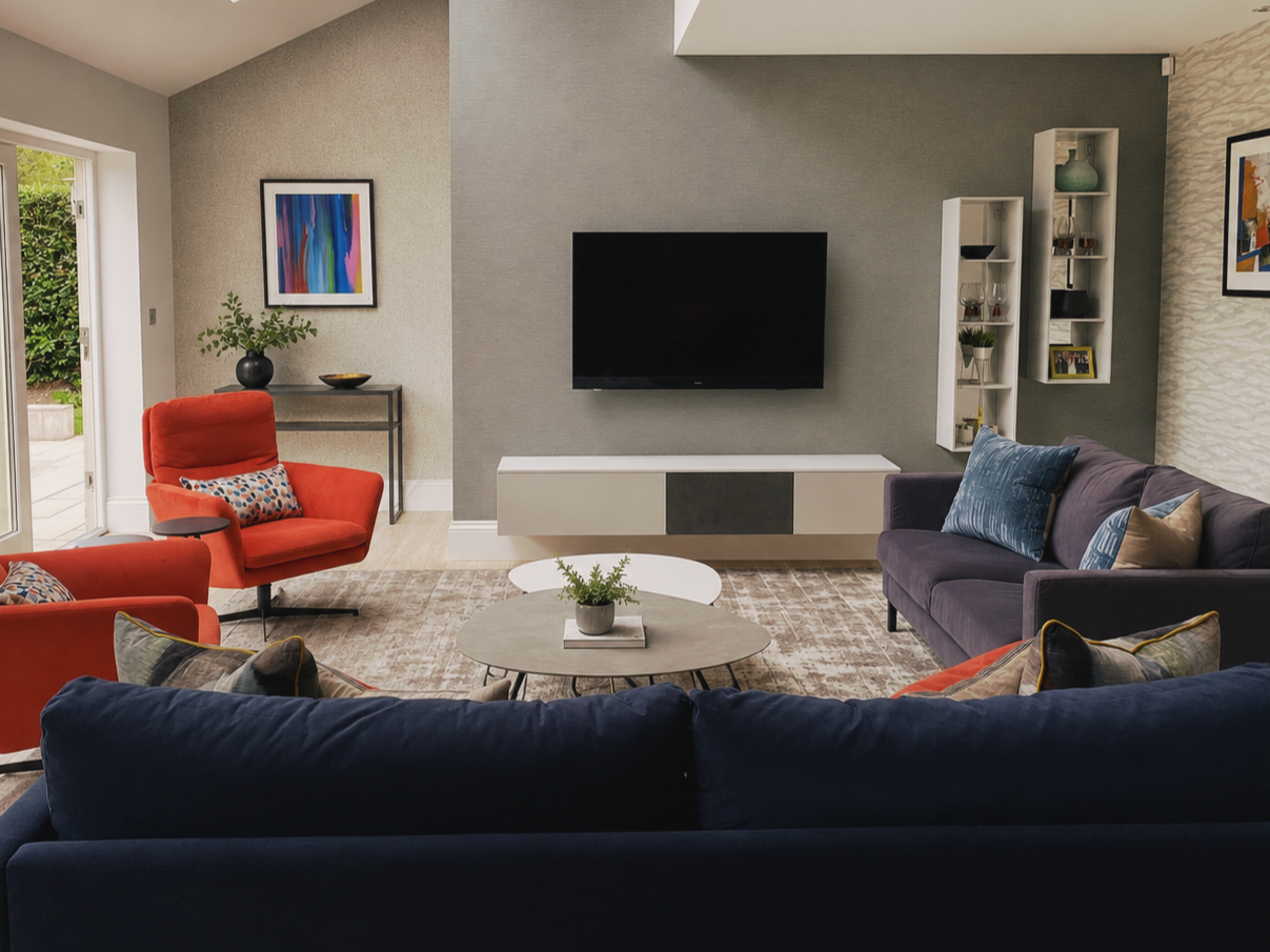 Modern living room with a black wall-mounted TV, gray and purple couches, two red armchairs, a round coffee table with a small plant, artwork on the walls, a side table with a plant, and a sliding glass door leading outside.