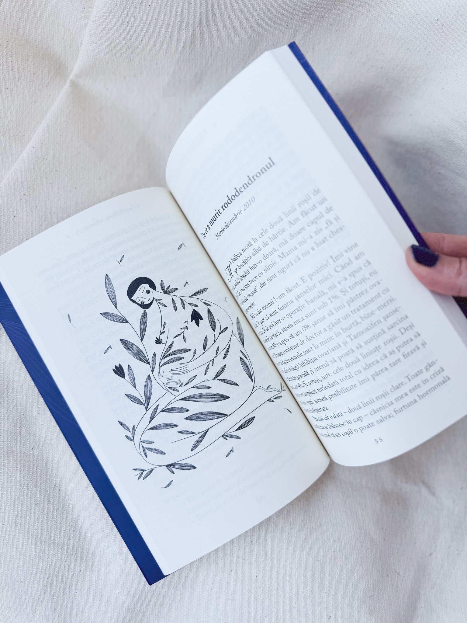 Open book with illustration of a mermaid surrounded by leaves on the left page and text in a foreign language on the right page, held by a person with dark painted nails.