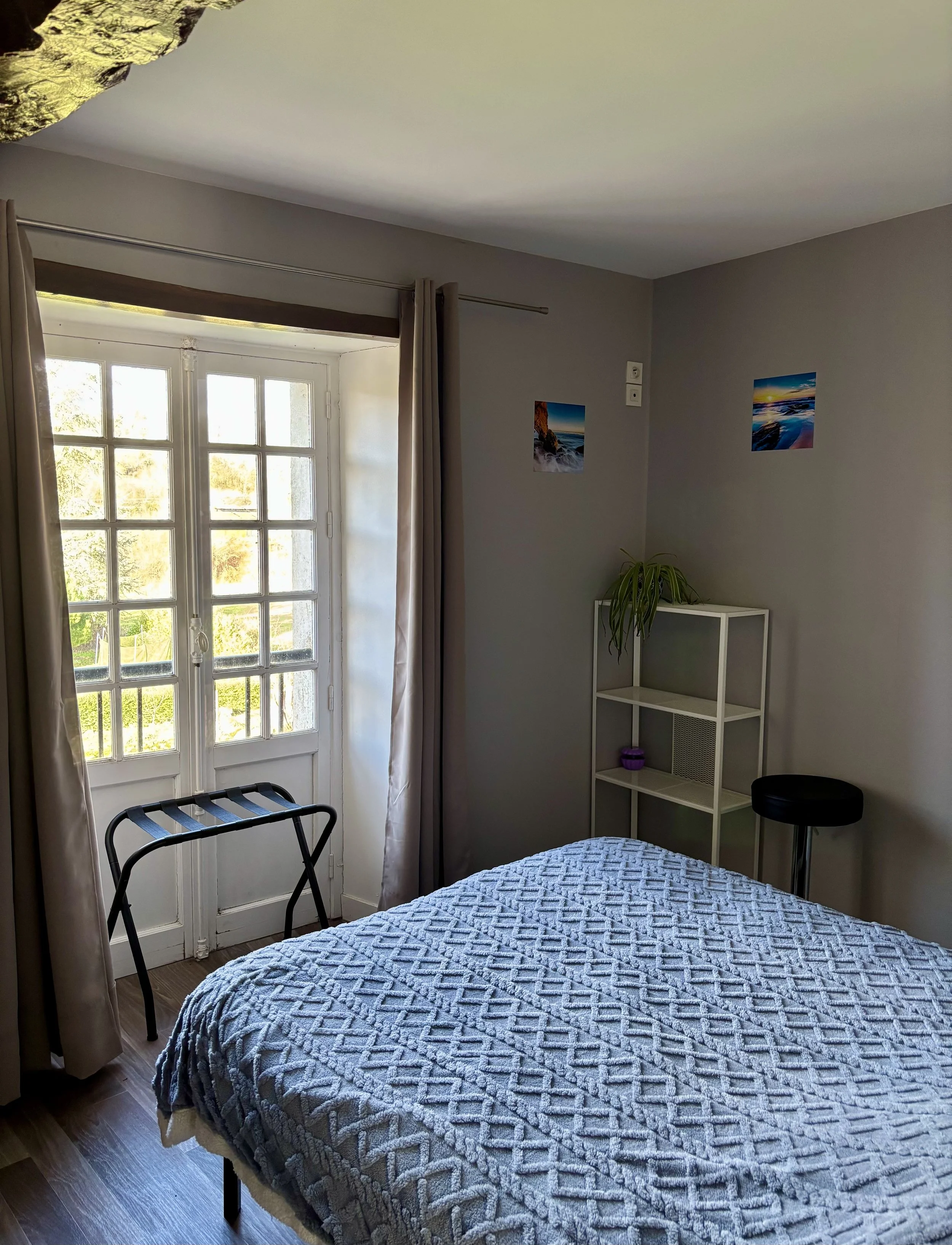 Chambre avec lit, fenêtre avec rideaux, étagère blanche avec une plante, tabouret noir, deux photos de paysage sur le mur.