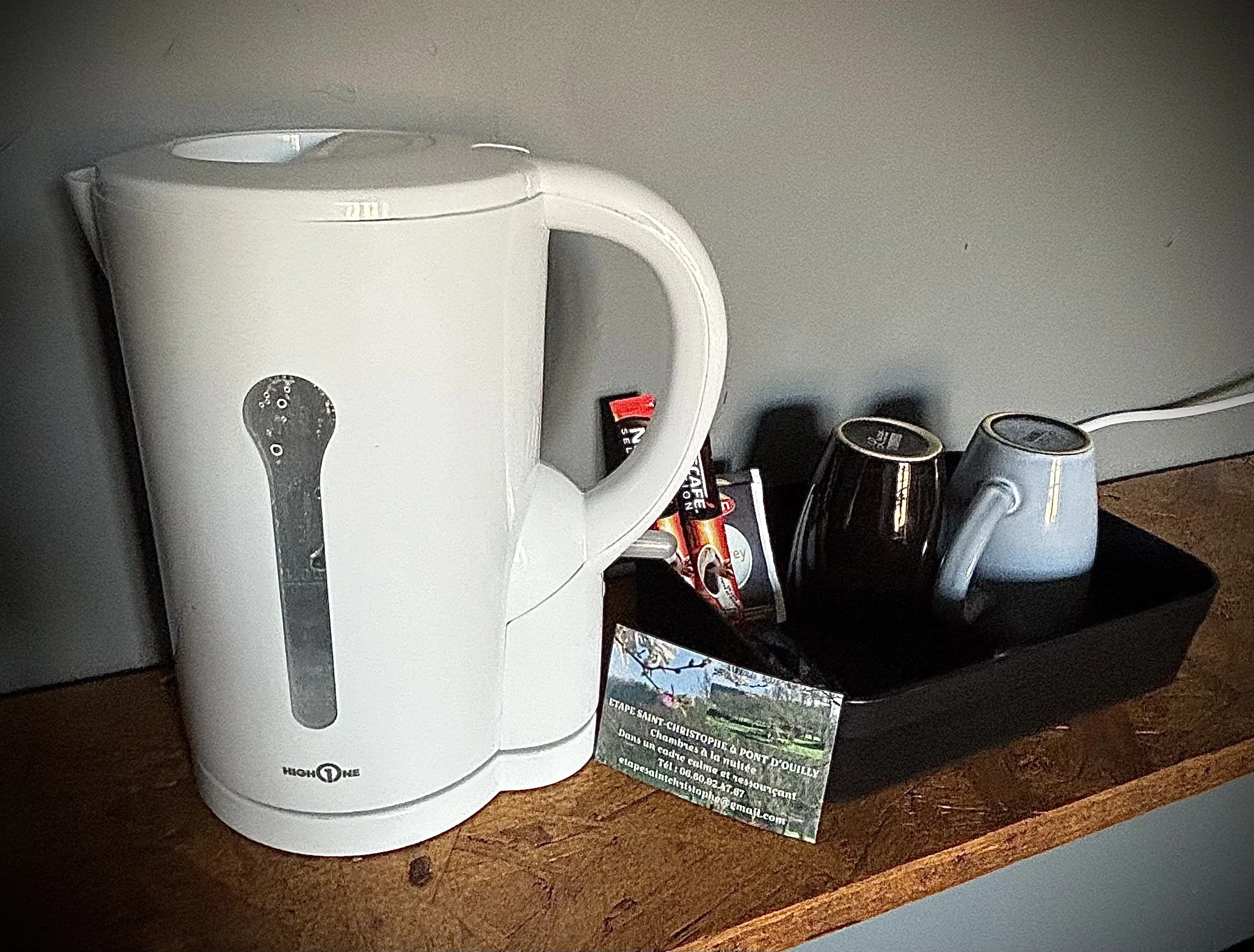 Une bouilloire électrique blanche, deux tasses (une noire et une blanche), deux sachets de thé ou café, et une carte de visite sur une surface en bois