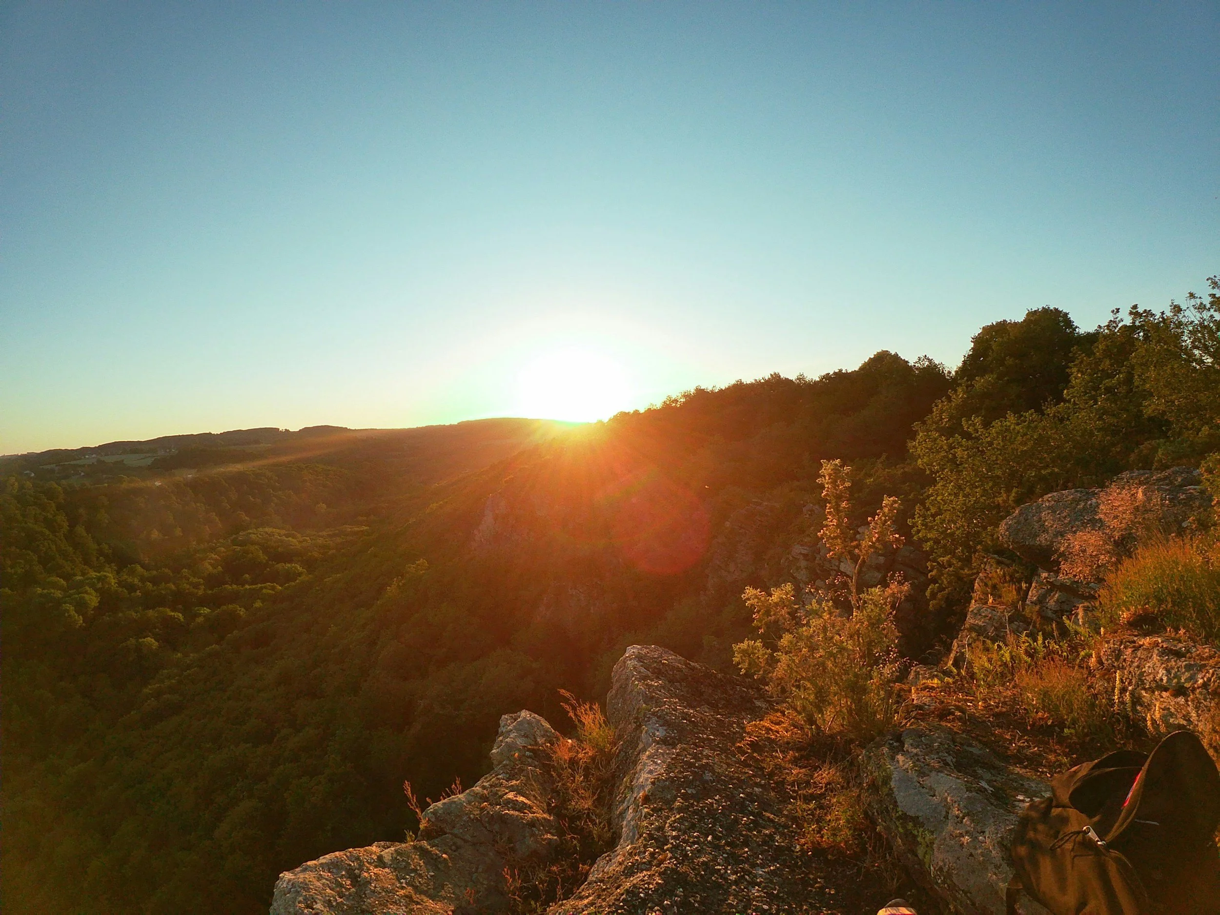 Coucher de soleil sur un paysage de collines boisées vues depuis une falaise avec des rochers et un sac à dos à proximité.