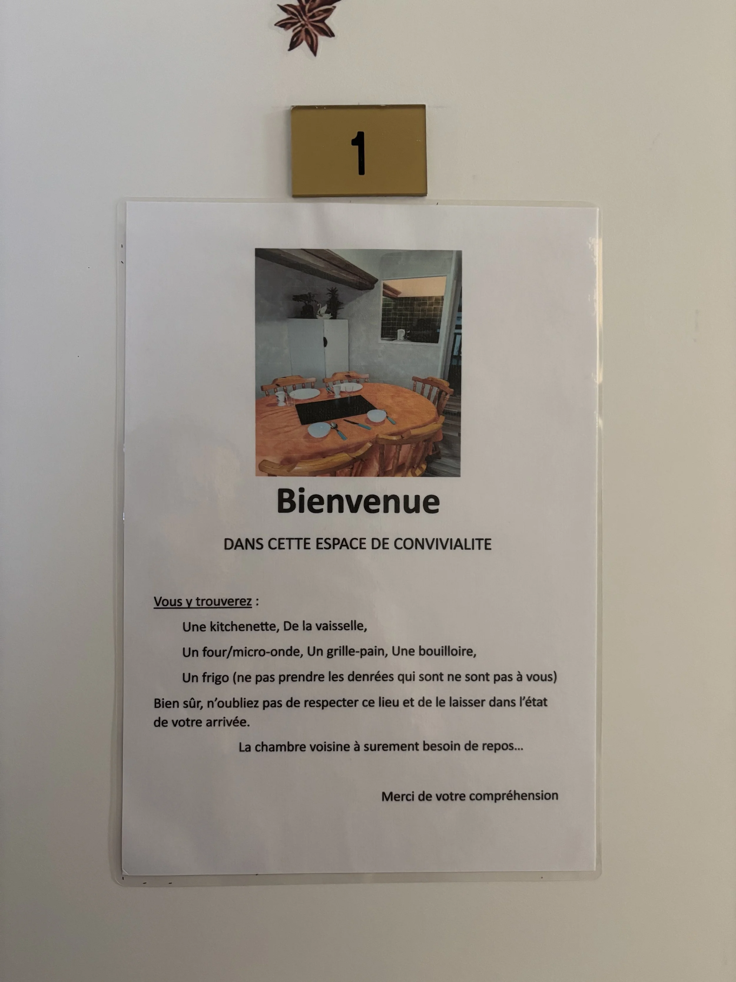 Une affiche de bienvenue placée au-dessus d'une porte, montrant une image d'une salle à manger avec une grande table en bois, entourée de chaises, et une pièce en arrière-plan. Le texte remercie les visiteurs et décrit les équipements disponibles dans l'espace.