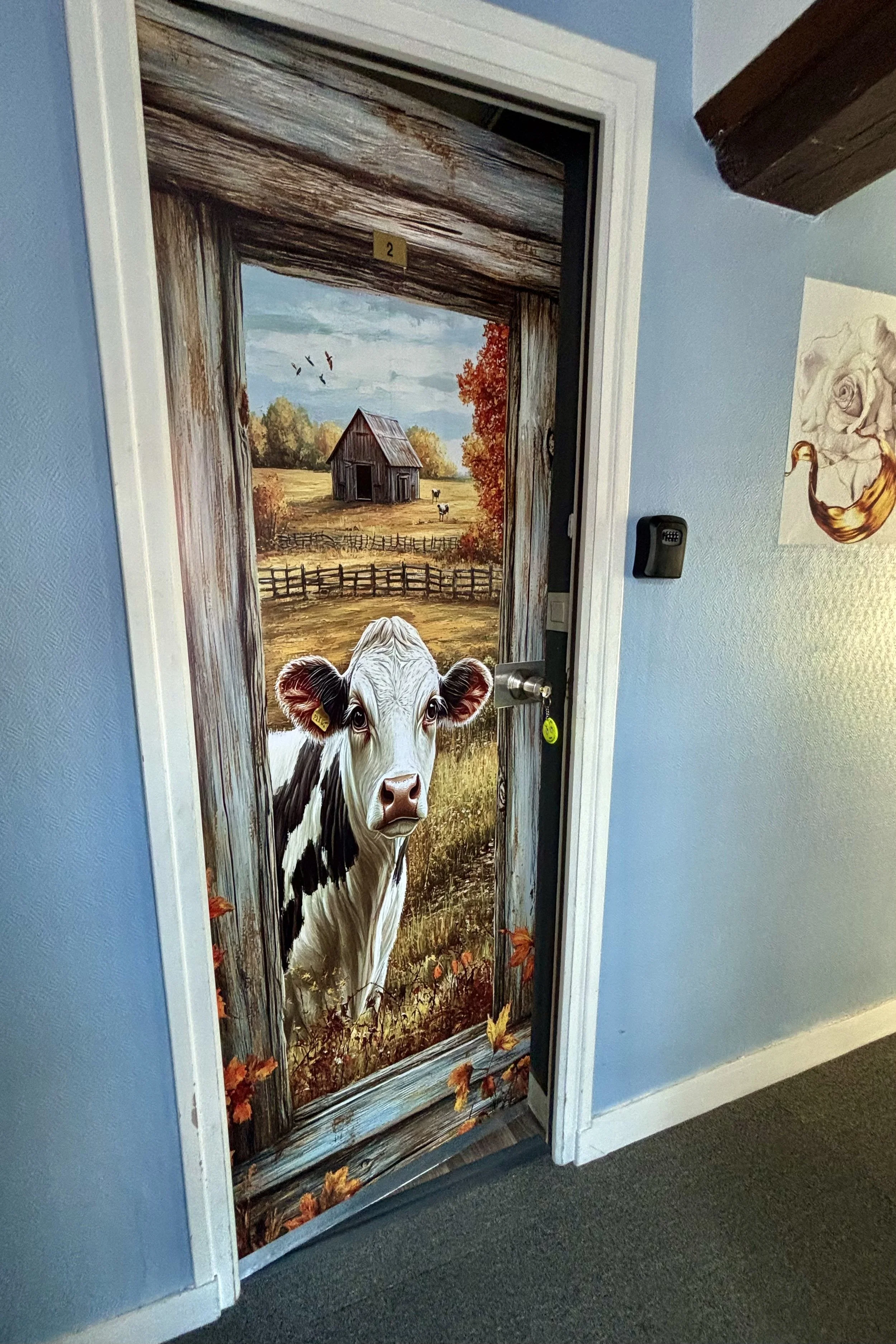Une porte peinte avec une scène de ferme représentant un veau regardant à travers une ouverture dans un cadre en bois, avec un paysage rural en arrière-plan.