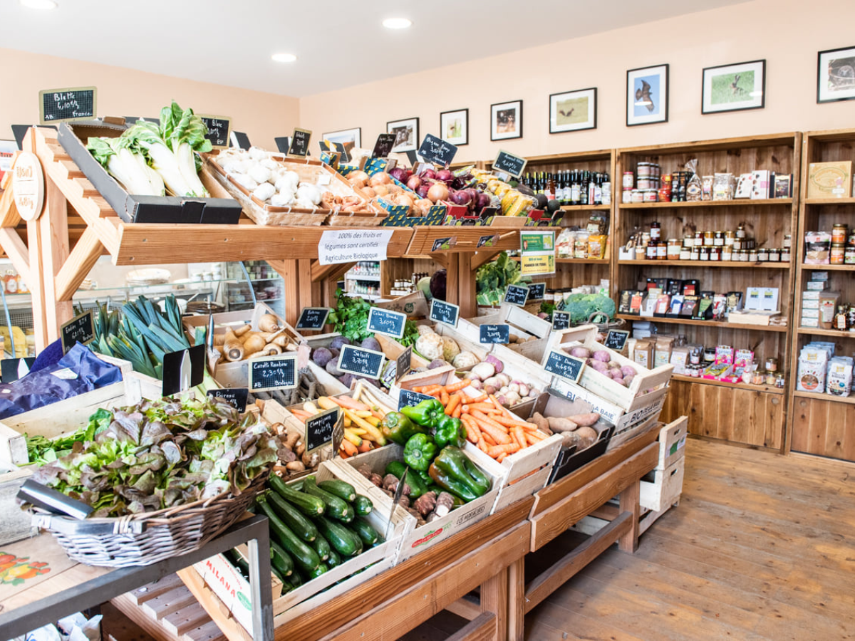 Marché de fruits et légumes bio avec différentes variétés de légumes frais, tels que courgettes, haricots verts, carottes, poivrons, oignons, ail, et autres, dans un magasin. Il y a aussi des épices et des produits en pots sur les étagères en bois derrière.