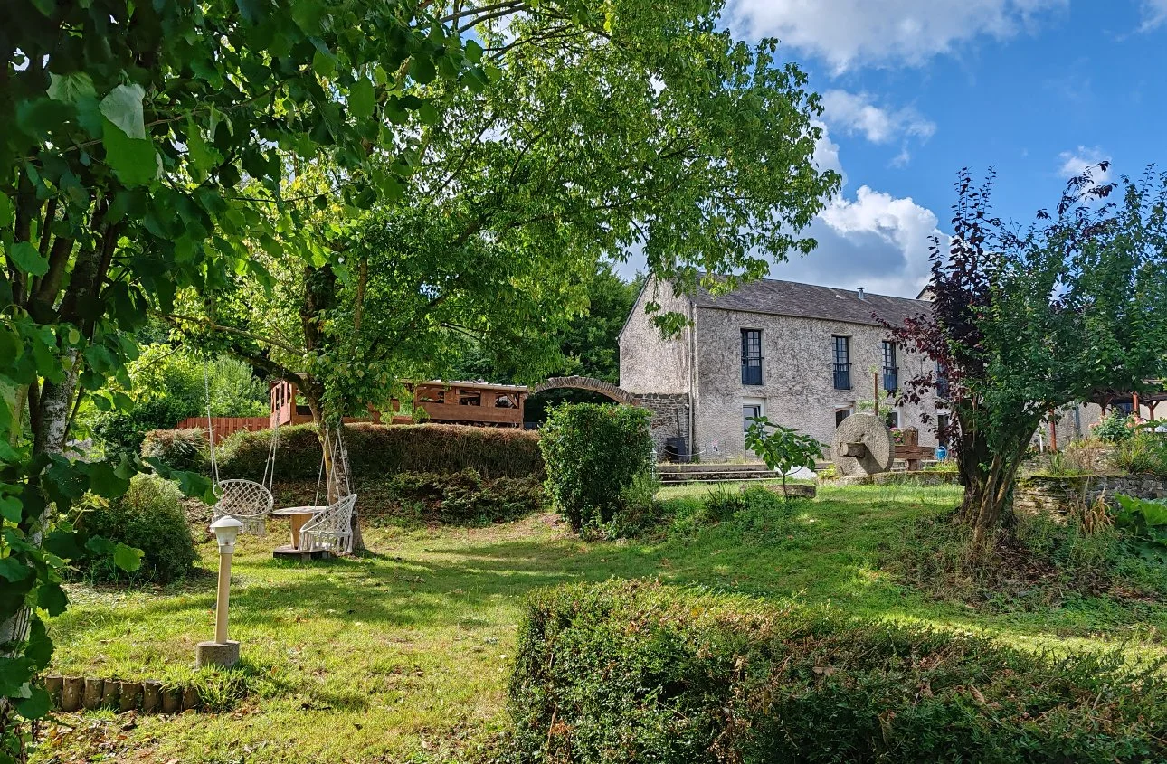 Un jardin verdoyant avec des arbres, un fauteuil suspendu en bois, une vieille maison en pierre, et un ciel bleu avec quelques nuages.