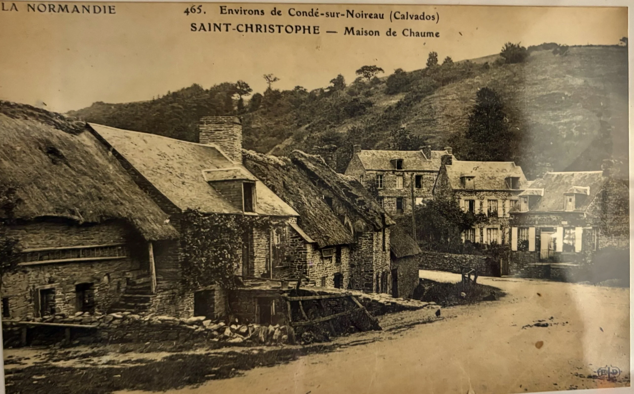 Photo en noir et blanc du village de Saint Christophe avec des maisons en pierre, des toits en chaume, des collines en arrière-plan, et une route non pavée passant devant les maisons, datant probablement du début du XXe siècle.