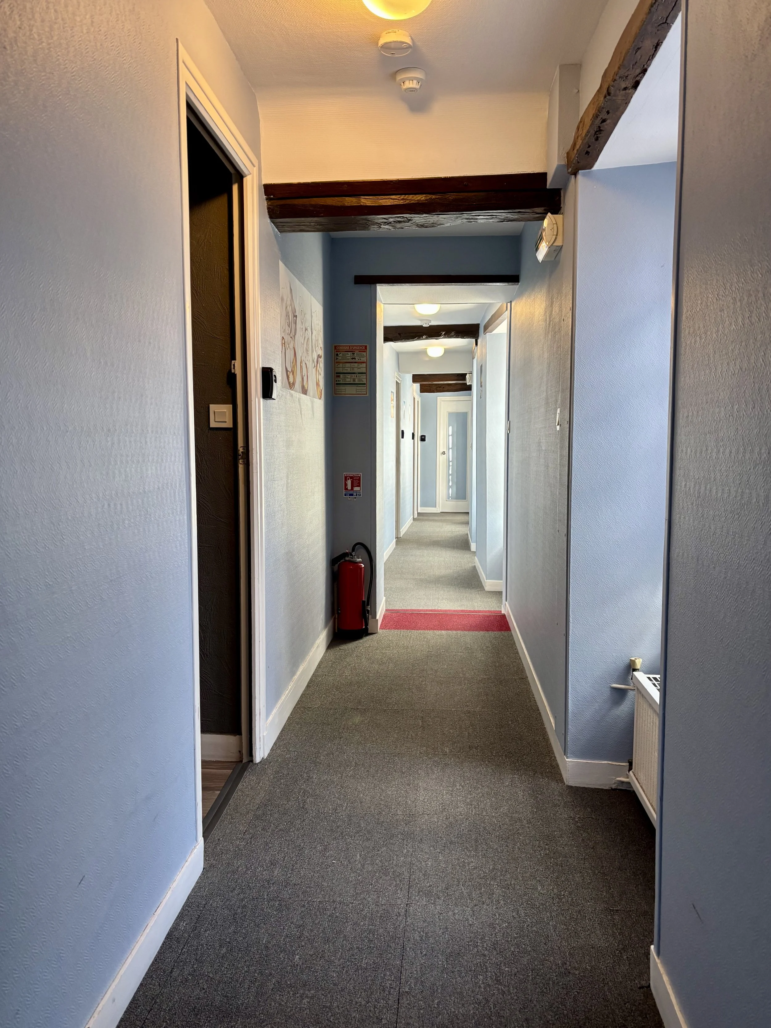 Couloir intérieur d'un bâtiment avec murs bleus, plafonds blancs, poutres en bois apparentes, et un tapis gris. Il y a une sortie de porte au fond et un extincteur rouge à gauche.