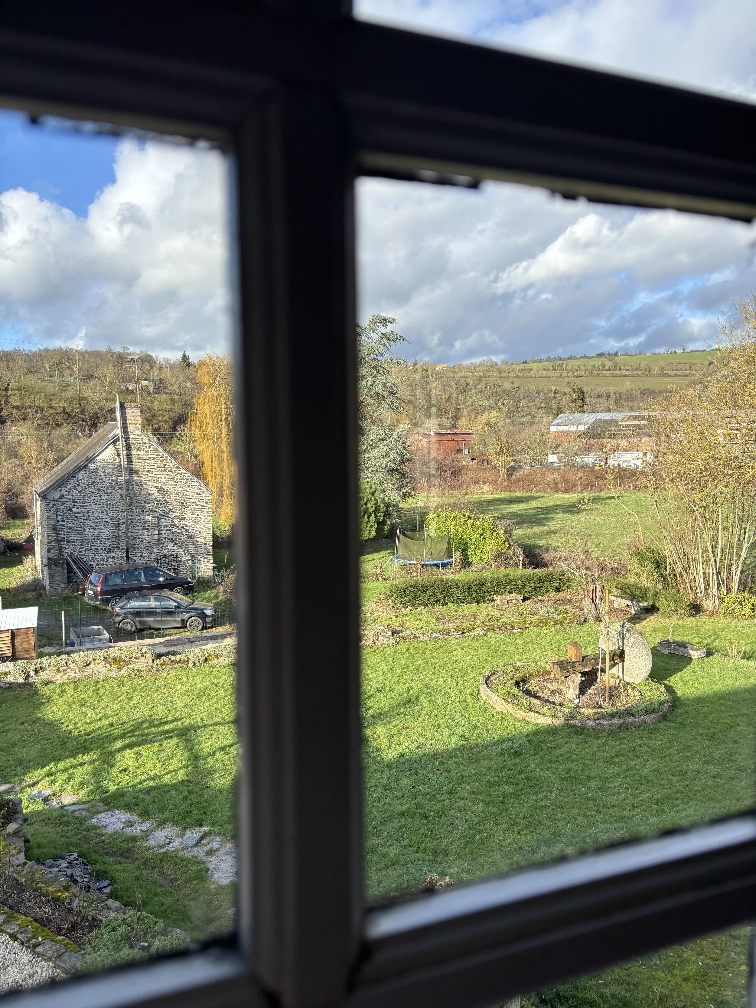 vue d'une cour extérieure avec un jardin, arbres, petites structures, et une maison en pierre, vue à travers une fenêtre.