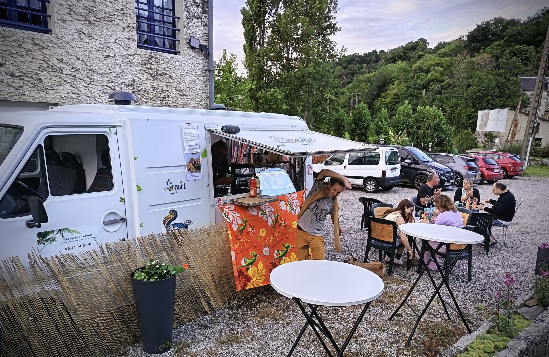 Un food truck blanc avec des décorations de flamants roses et une toile orange, vendant de la nourriture dans une zone extérieure, avec des personnes assises à une table à manger et un homme debout à côté du food truck. En arrière-plan, des voitures sont garées devant une forêt verdoyante.