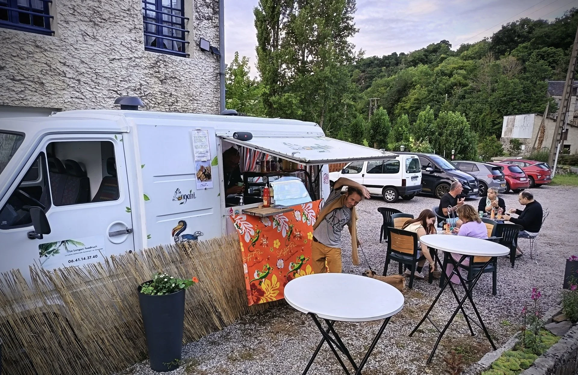 Un food truck blanc avec des décorations de toucan et de feuilles, où un homme se tient près d'une table avec un parasol orange, et un groupe de personnes dine à une table en extérieur avec plusieurs autres véhicules en arrière-plan, dans un environnement naturel avec des arbres et des collines.