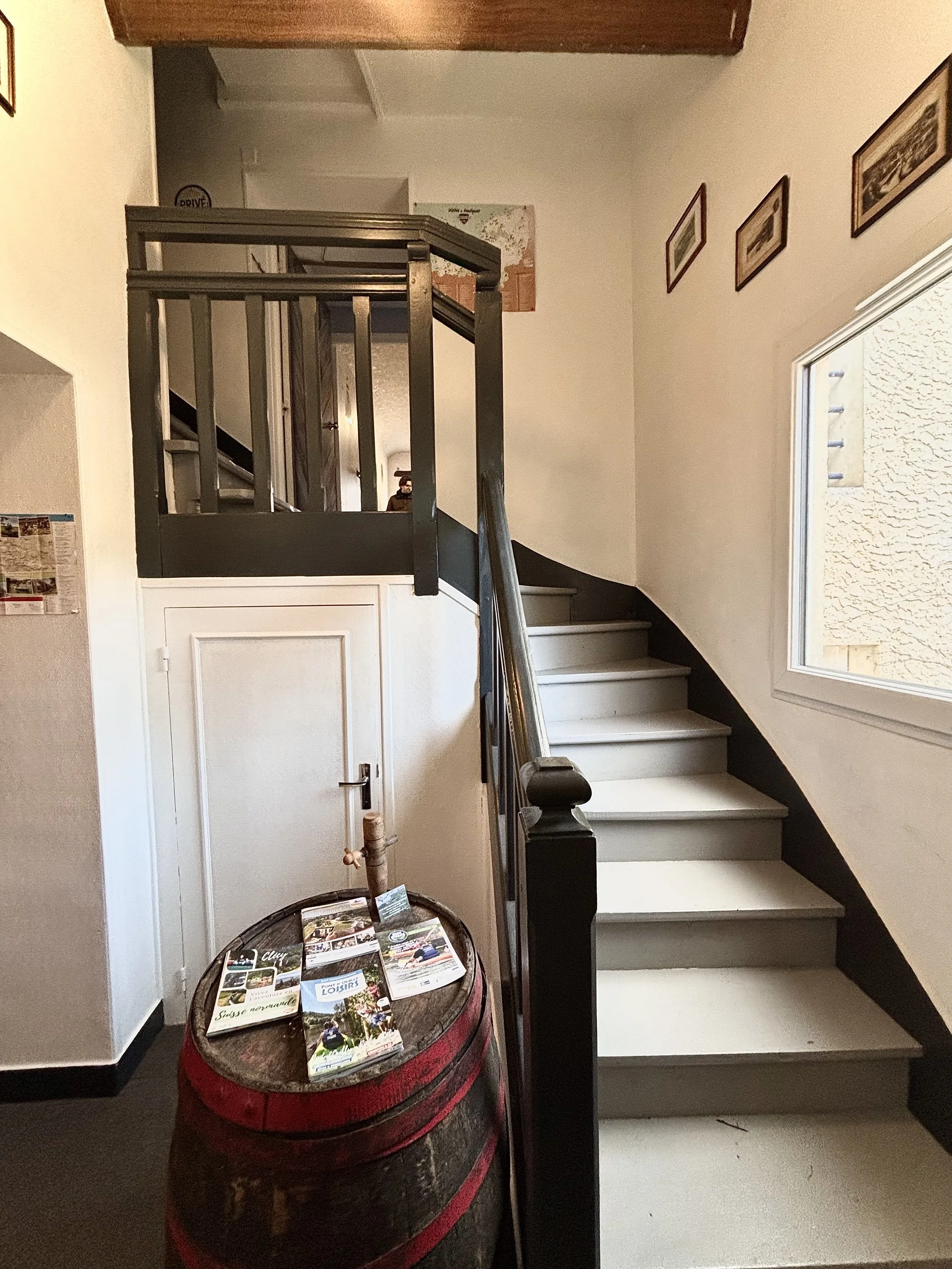 Escalier de bois avec rampe noire dans un intérieur, avec un mur blanc, une fenêtre et des cadres accrochés, et un tonneau de whisky recyclé utilisé comme table avec des brochures de la région, suisse normande, dessus.