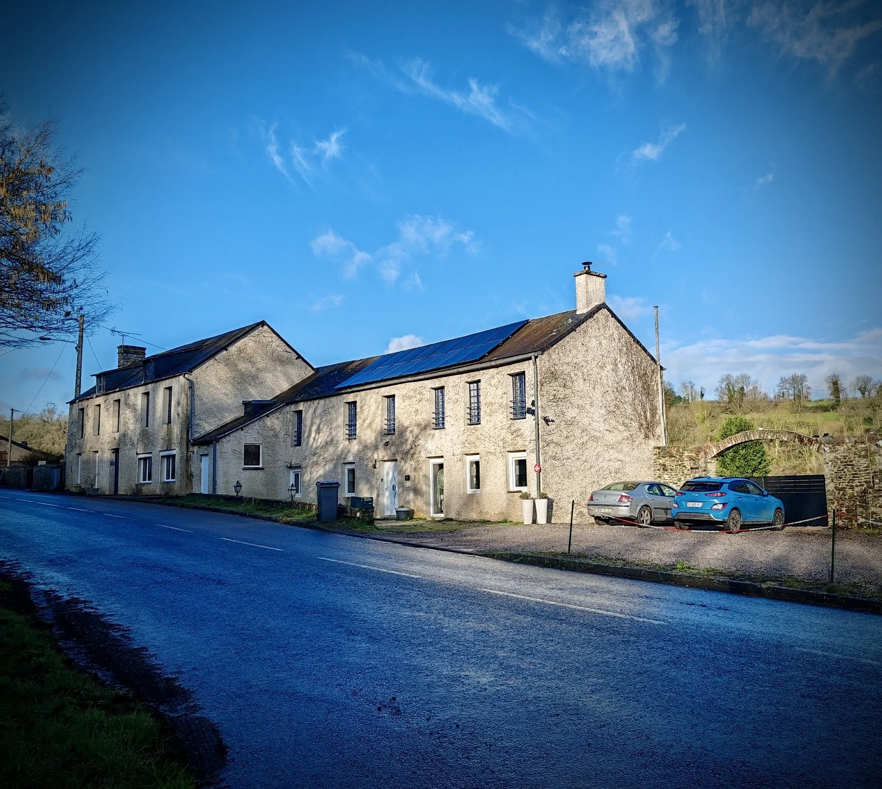 Maisons en pierre sous un ciel bleu avec des voitures garées devant, vue de rue dans un village rural.