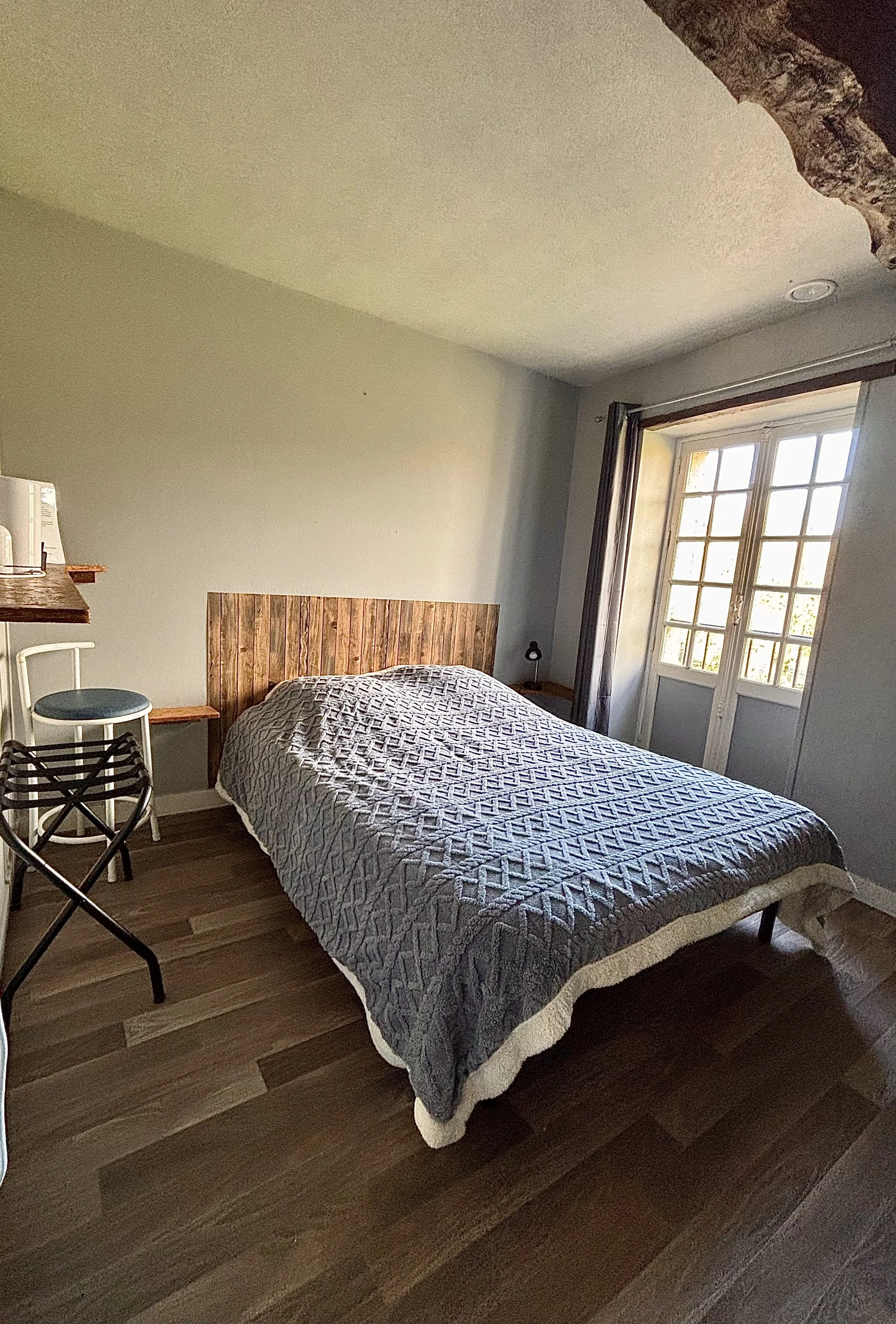 Chambre avec un lit, une tête de lit en bois, une fenêtre avec des volets, un tapis en paille, et un plafond avec une poutre en bois.