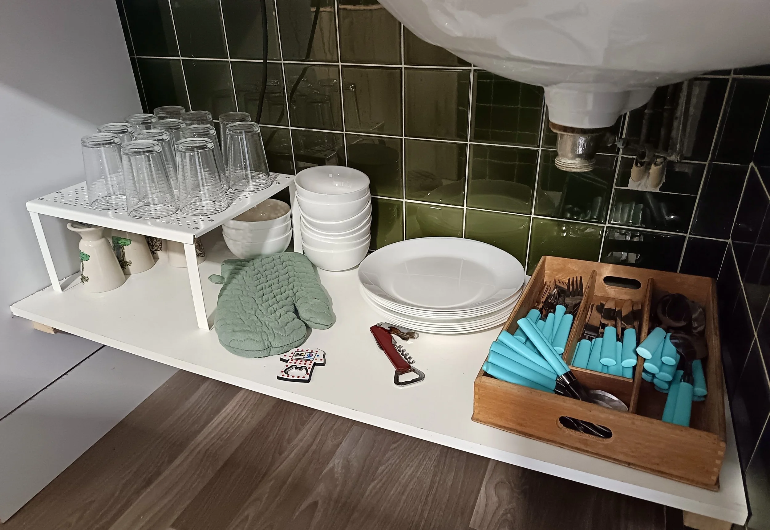 Vitrine de vaisselle dans une cuisine, avec des verres, des bols, des assiettes, des couverts, des rouleaux de film plastique, un décapsuleur, et des gants en tissu vert, le tout organisé sur une table blanche avec un fond en carreaux vert foncé.