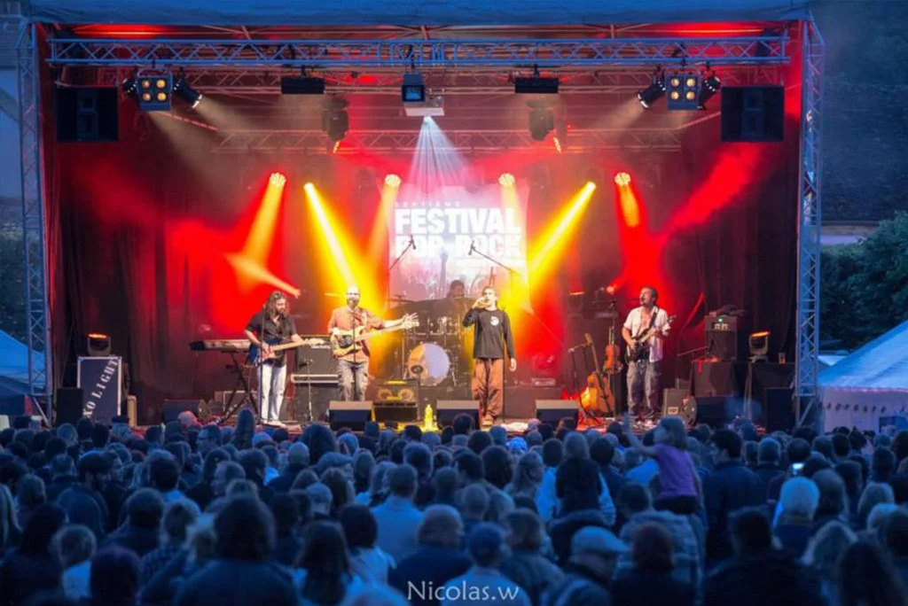 Un groupe de musiciens joue sur scène lors d'un festival en plein air, avec un grand public devant et un affichage lumineux en arrière-plan.