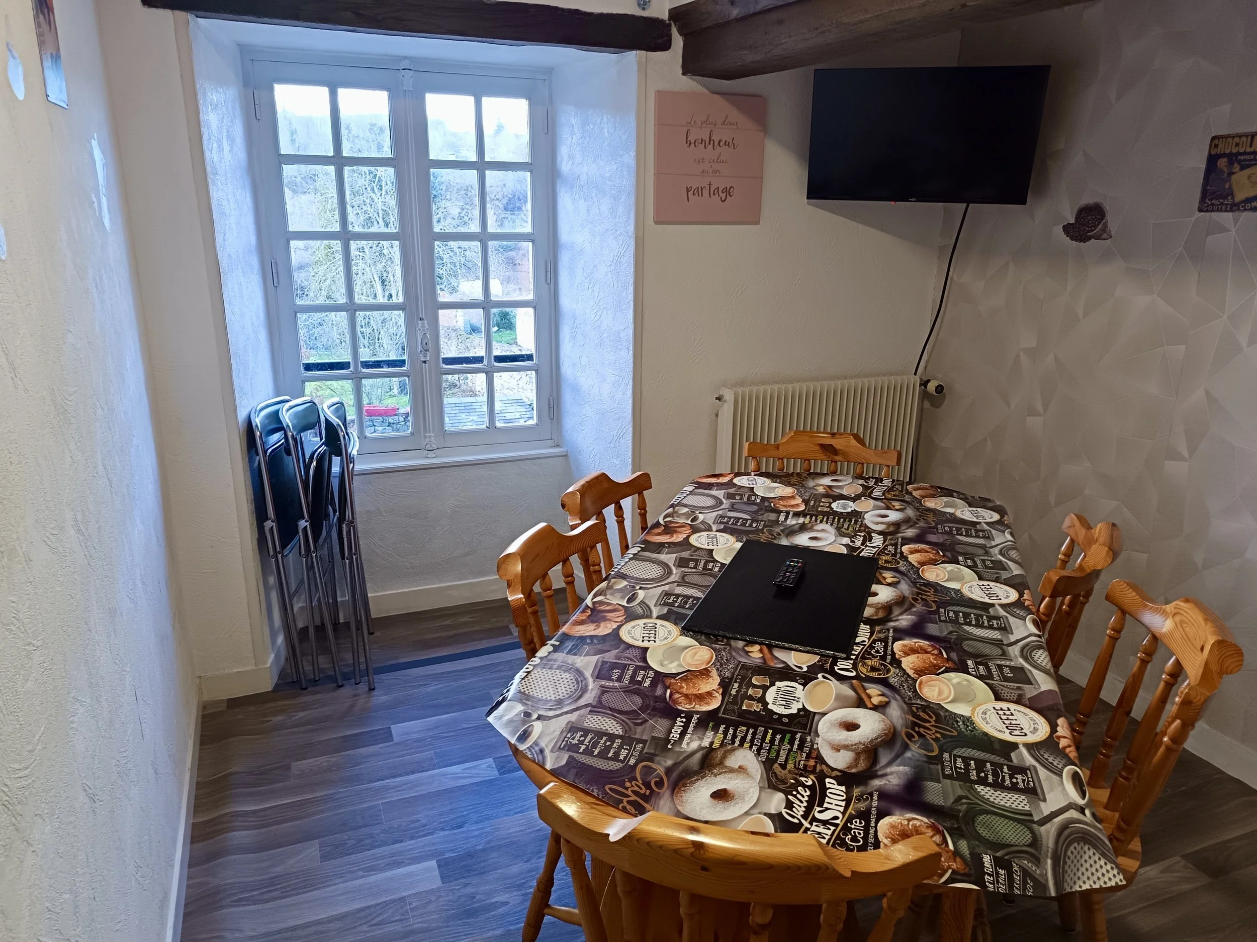 Salle à manger avec une table recouverte d'une nappe à motif de donuts, entourée de six chaises en bois. À gauche, une fenêtre laissant entrer de la lumière. Sur le mur derrière la table, un radiateur et une télévision suspendue. À droite, un poster rose avec un texte en français et plusieurs petits objets.