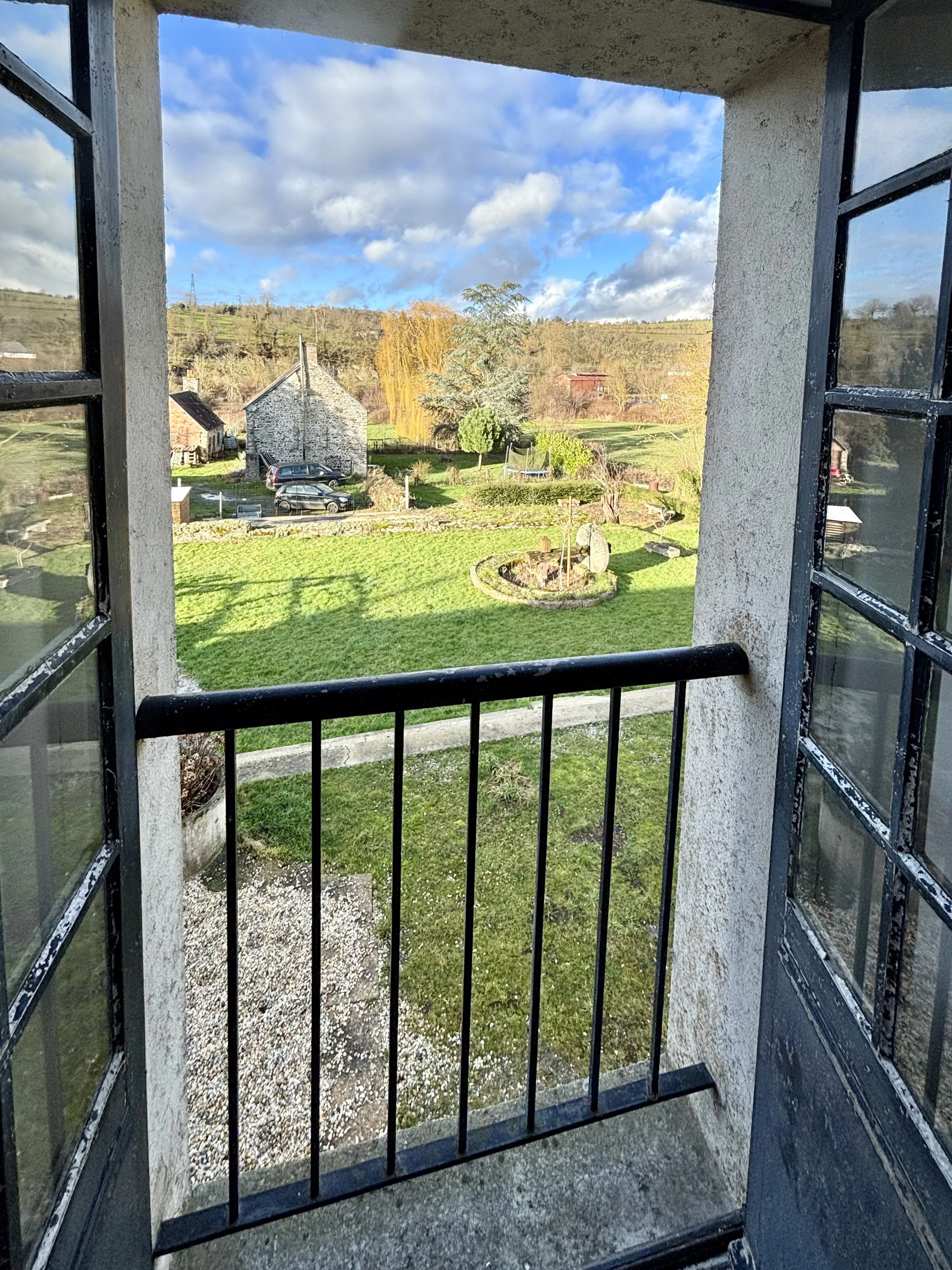 Vue d'un jardin verdoyant depuis une petite balustrade de fenêtre en bois, avec un ciel bleu parsemé de nuages, et des maisons et arbres en arrière-plan.