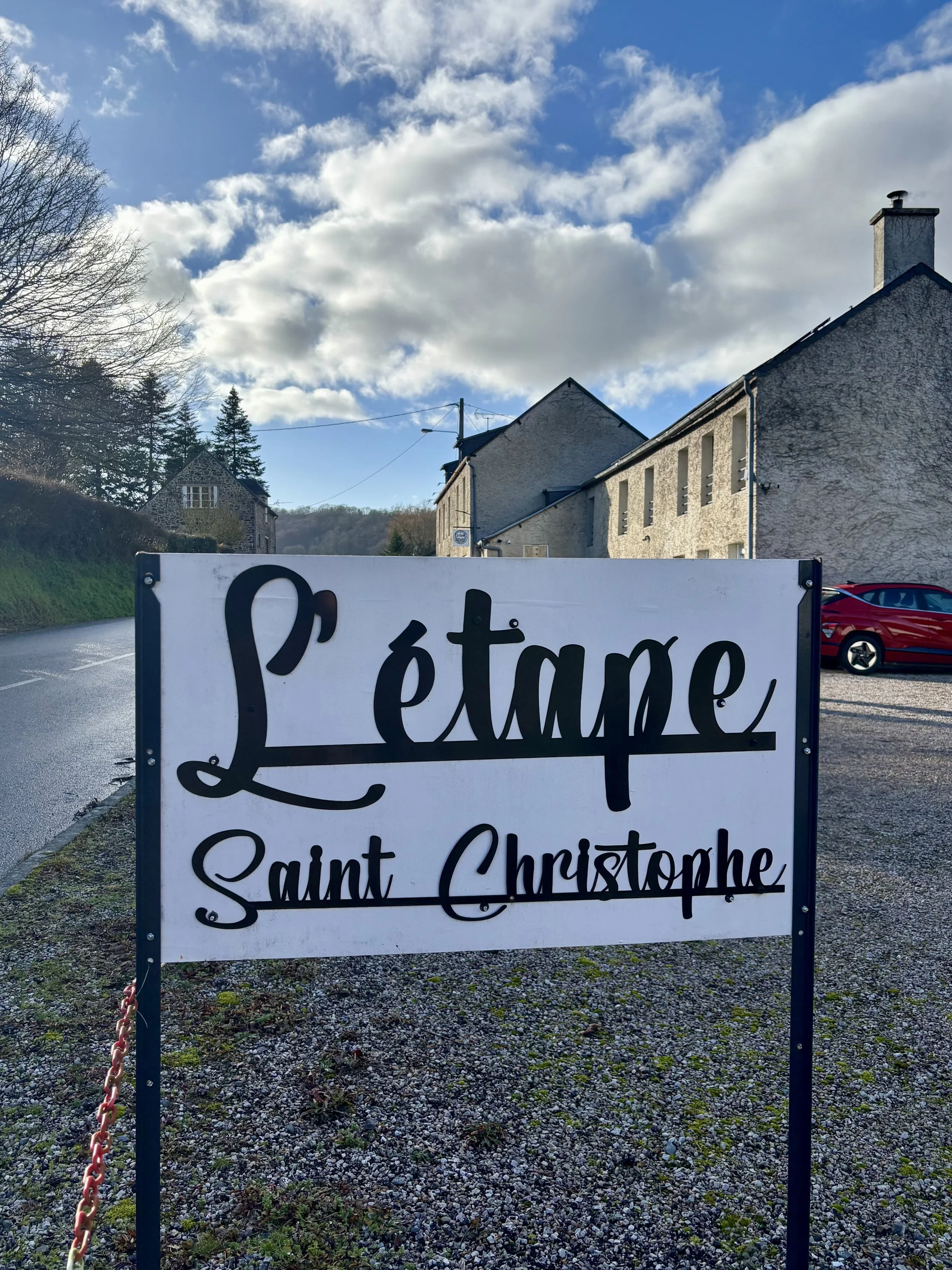 Panneau indiquant 'L'étape Saint Christophe' à côté d'une rue dans un village avec des maisons anciennes en pierre et un ciel partiellement nuageux.