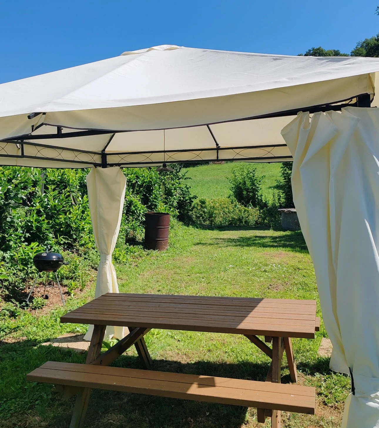 Un gazebo blanc avec un toit en toile blanche, une table en bois et un banc en bois dans un jardin ensoleillé avec de la verdure et un barbecue noir à gauche.