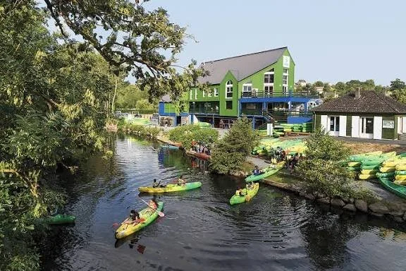 Une scène au bord d'une rivière avec plusieurs kayaks verts et des personnes qui pagayent, un grand bâtiment vert et bleu en arrière-plan, entouré d'arbres et de végétation.