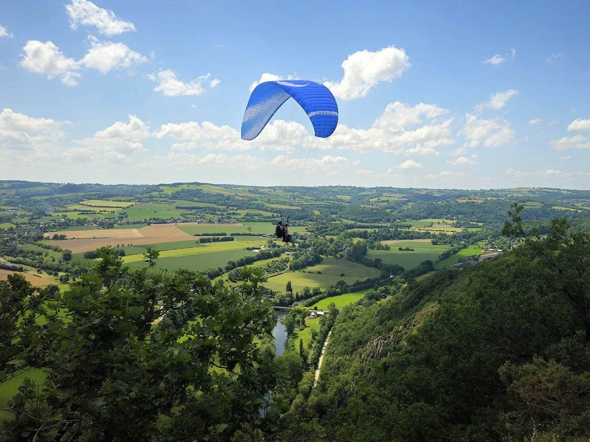 Personne faisant du parapente au-dessus de paysages ruraux avec des champs et des collines, ciel partiellement nuageux.