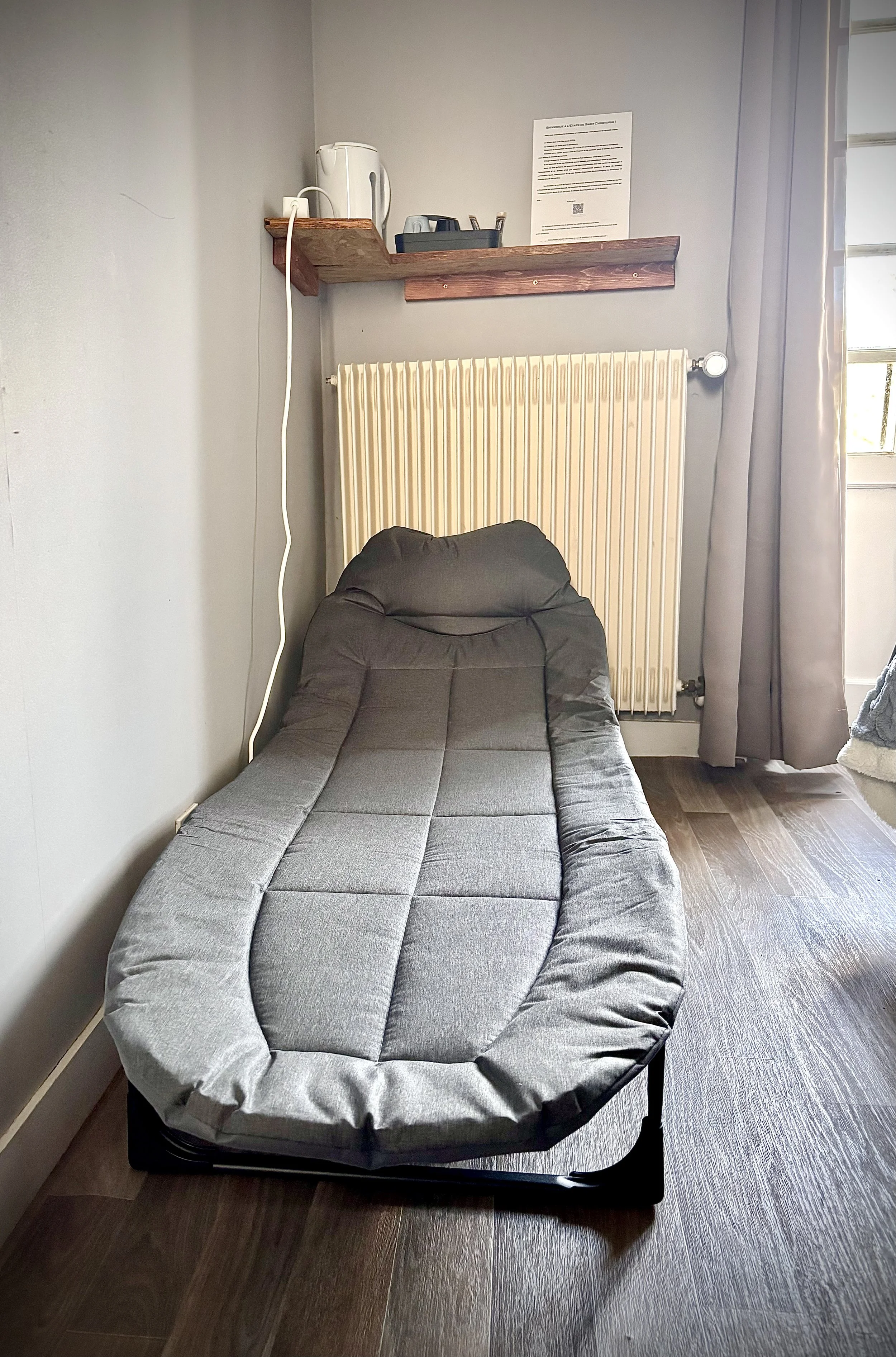 Une pièce avec un canapé-lit gris, un radiateur blanc, une étagère en bois avec une bouilloire électrique, une fiche de papier et un grille-pain, une fenêtre avec un rideau gris.