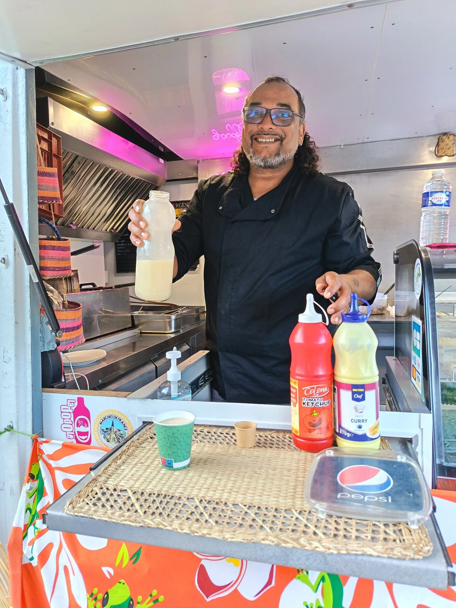 Un homme souriant dans un stand de nourriture, tenant une bouteille de lait ou de smoothie, avec des sauces ketchup et curry sur le comptoir devant lui.