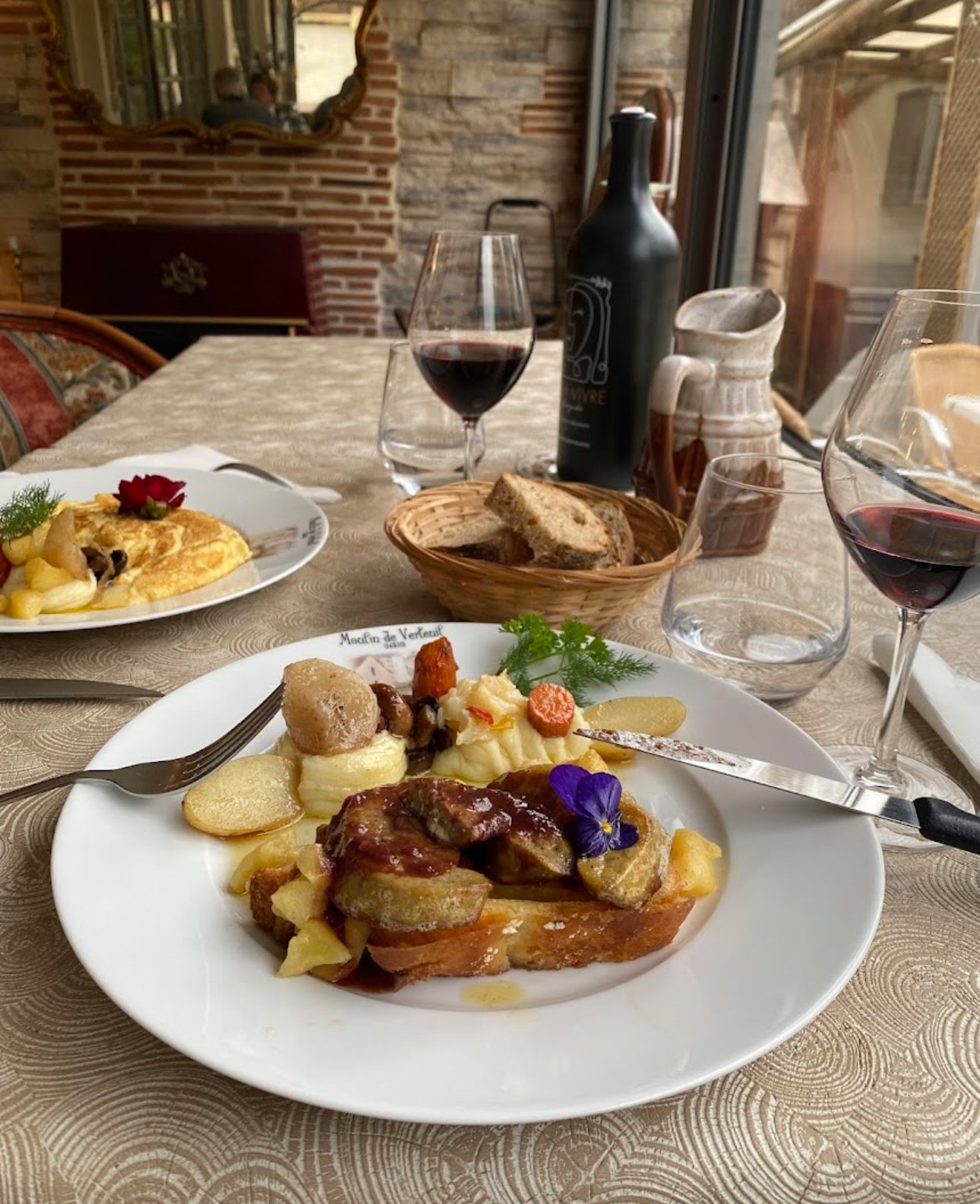 Repas français avec un plat principal de viande, pommes de terre et légumes, une assiette d'omelette, deux verres de vin rouge, du pain, et une carafe en arrière-plan dans un décor rustique.