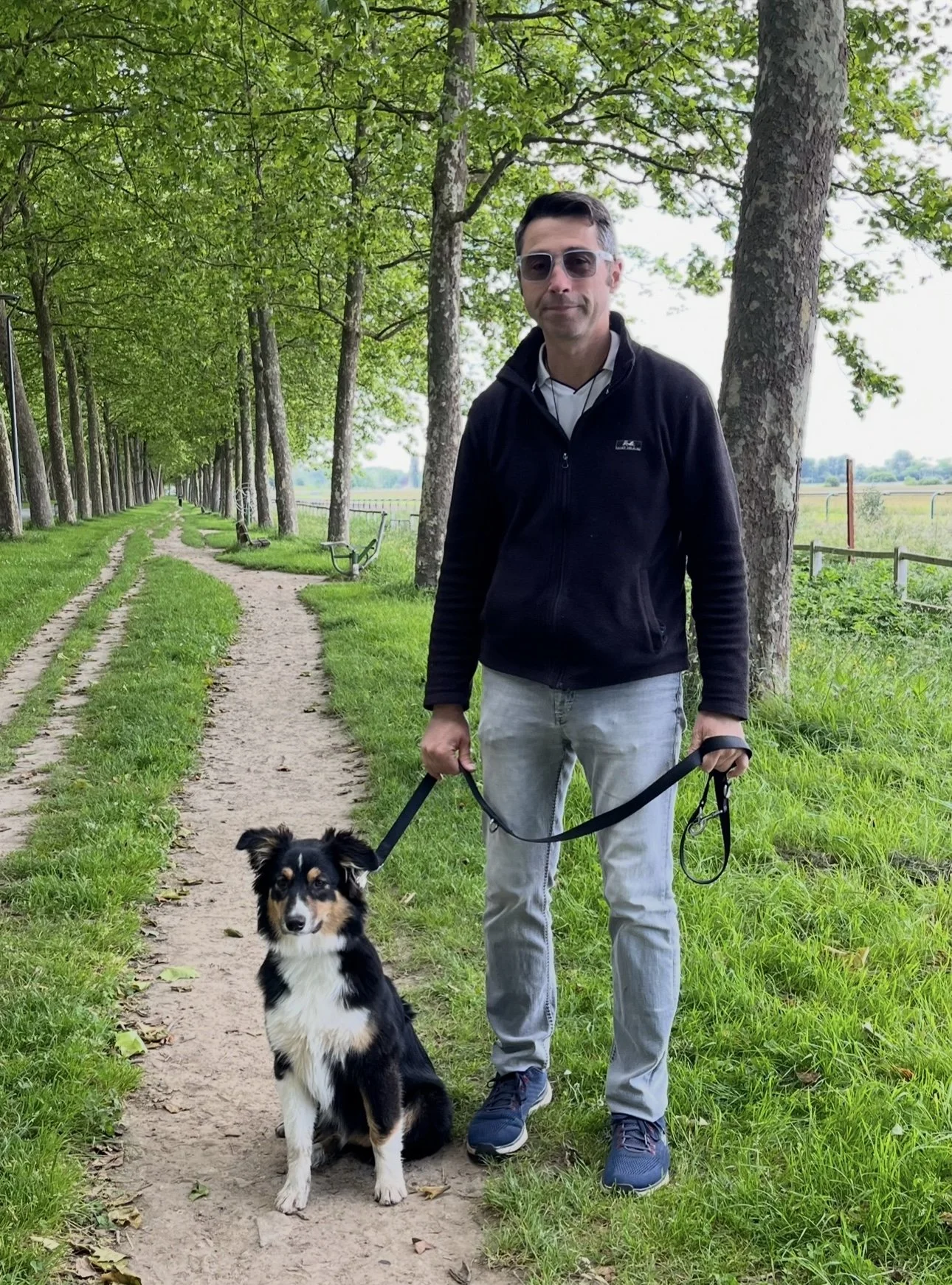 Florian Lebiez, homme avec des lunettes de soleil, portant une veste noire, tient la laisse d’un chien noir et blanc, assis sur un chemin forestier bordé d’arbres verts.