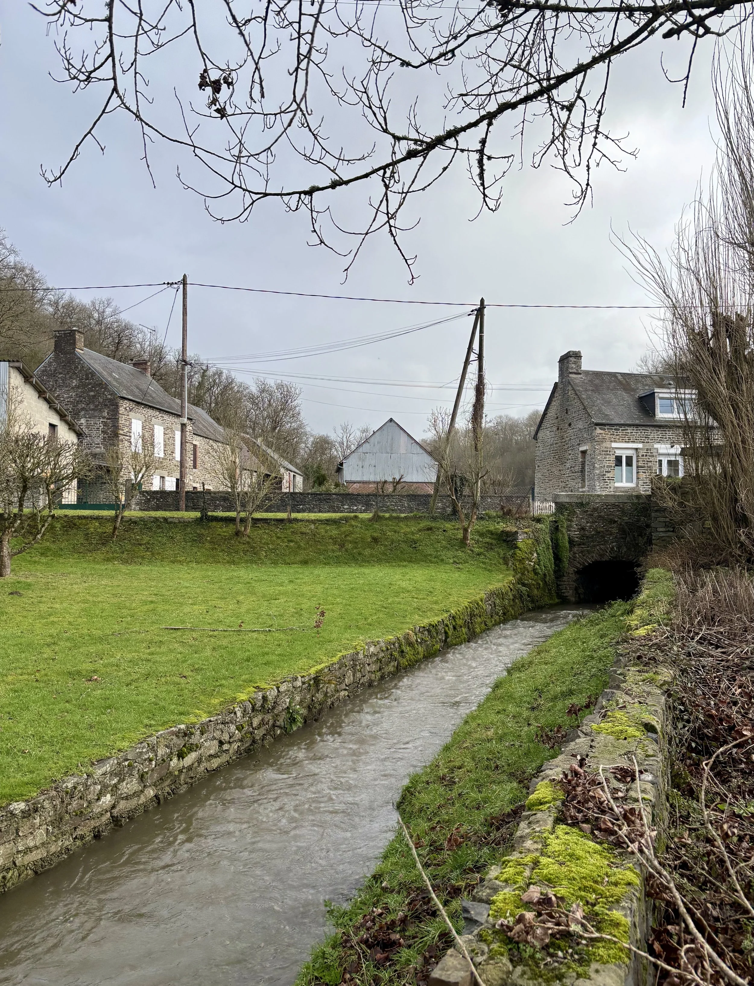 Petite rivière passant sous un pont en pierre, maisons en pierre en arrière-plan, arbres sans feuilles, ciel nuageux.