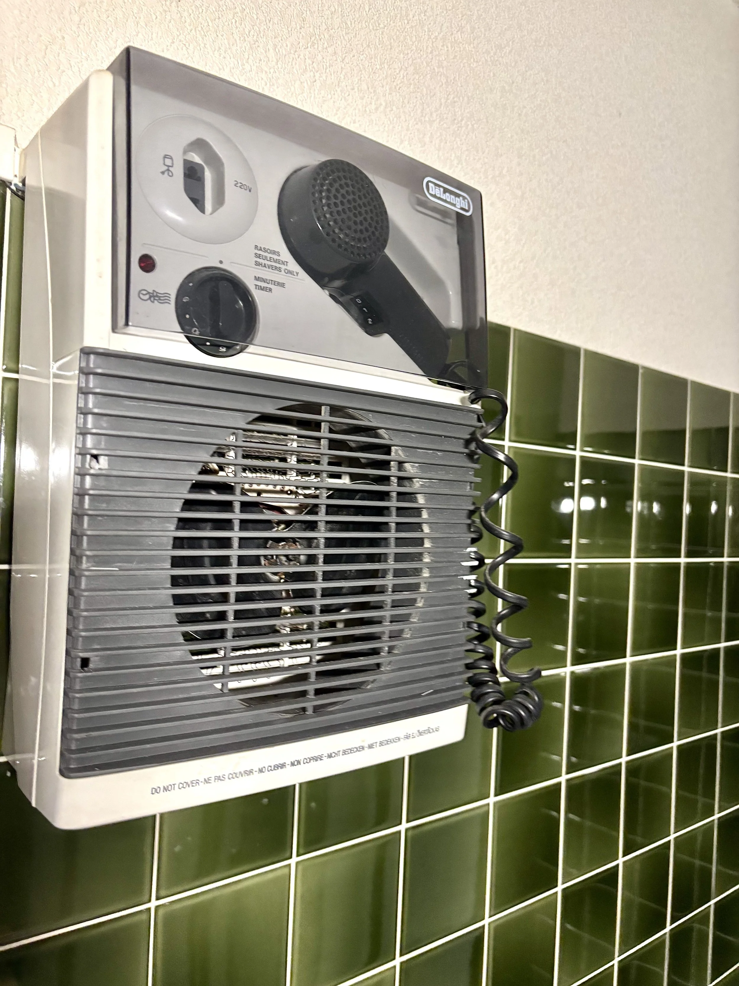 Tondeuse à cheveux avec capot noir et bouton noir, montée sur un mur avec carreaux verts en arrière-plan.