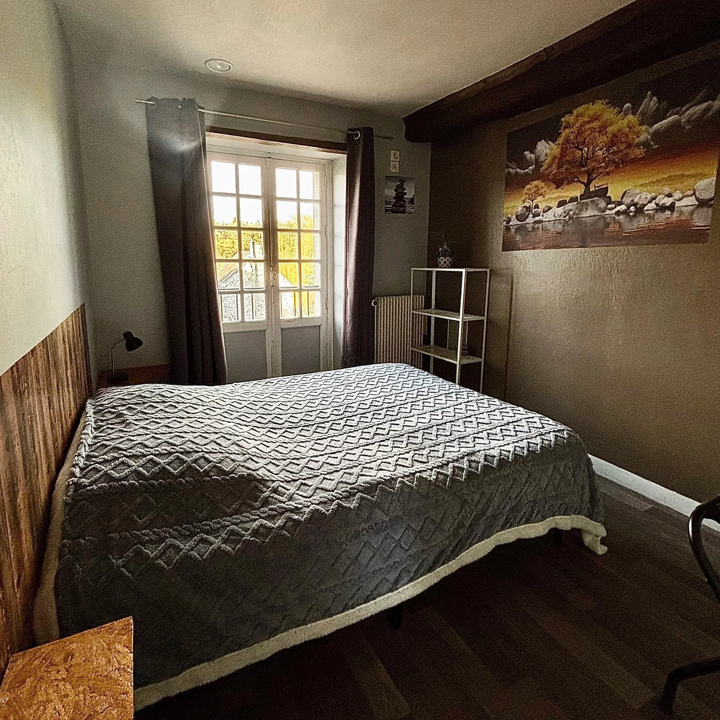 Chambre avec lit, vue sur une fenêtre, tableau d'un arbre sur la muraille, étagère et lampe de chevet.