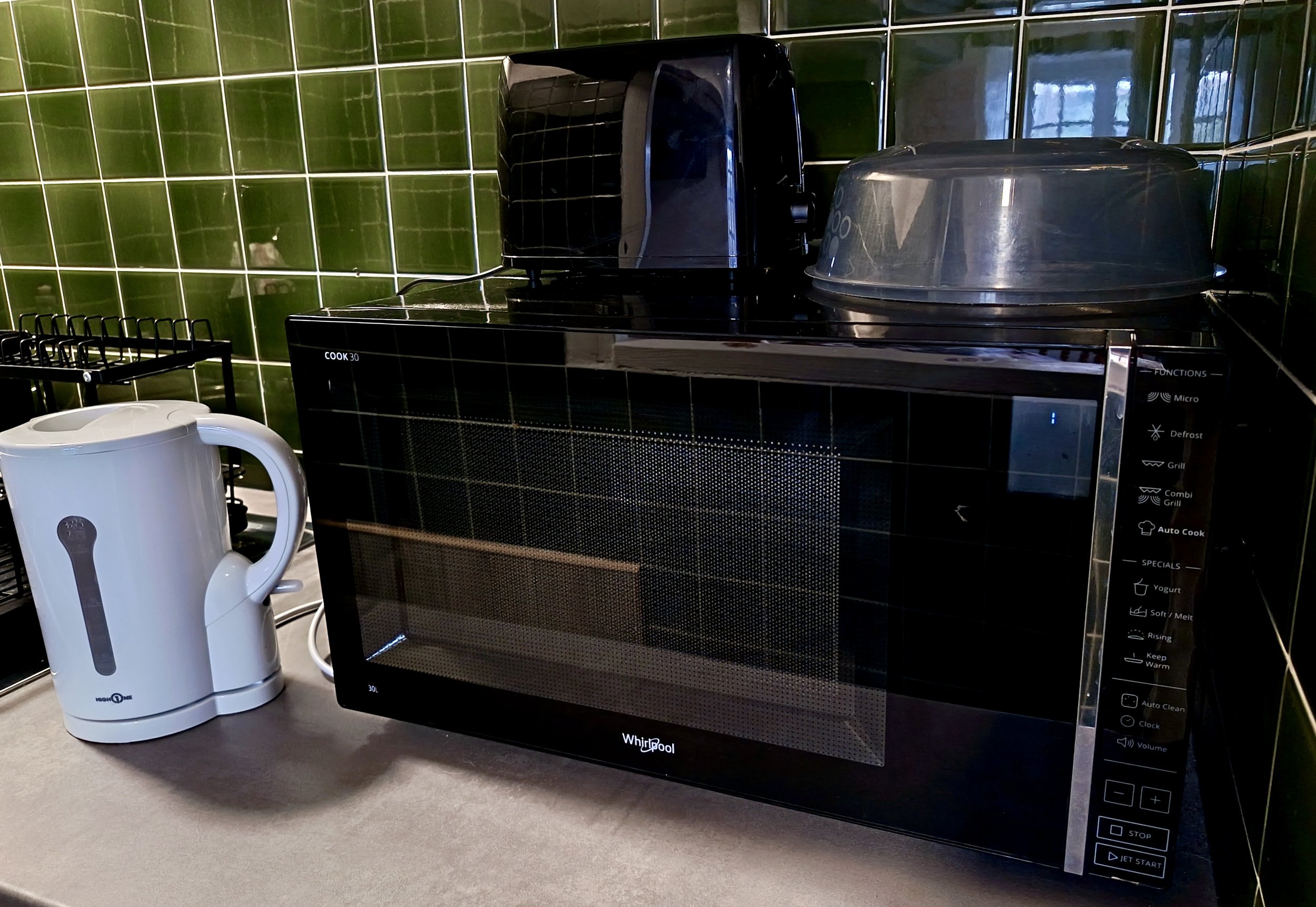 Four micro appliances: a white electric kettle, a black toaster, a microwave oven, and a black food steamer, on a kitchen countertop with green tiled walls.