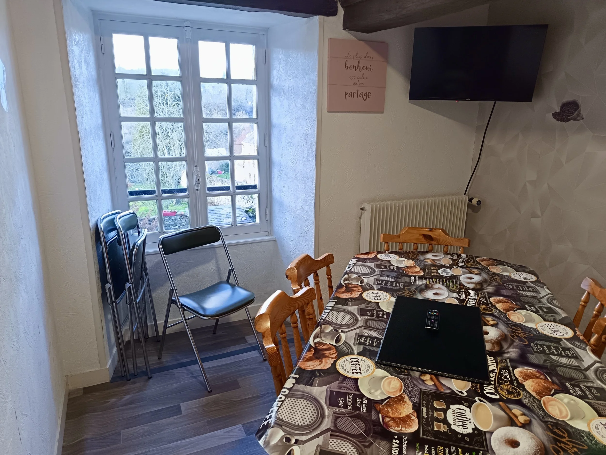 Salle à manger avec une table recouverte d'une nappe à motifs, des chaises en bois, un téléviseur fixé au mur, une fenêtre laissant passer la lumière naturelle, et une affiche sur le mur avec un message en français.