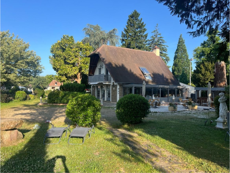 Maison à deux étages avec un toit en pente, grande terrasse ensoleillée, entourée d'arbres et de mobilier de jardin, sous un ciel bleu clair.