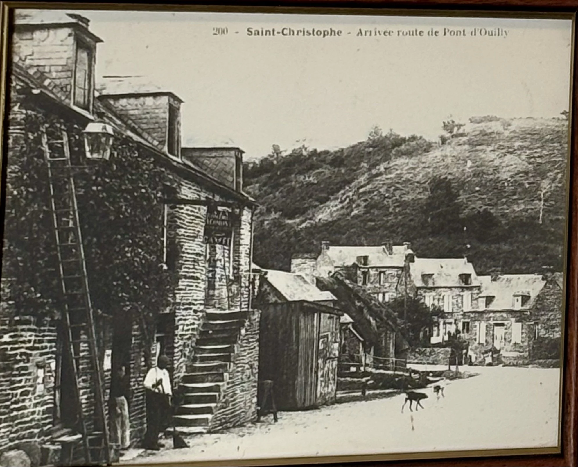 Photo en noir et blanc du village Saint Christophe, avec une colline boisée en arrière-plan. Titre en haut : 'Saint-Christophe - Arrivée route de Pont d'Ouilly'.
