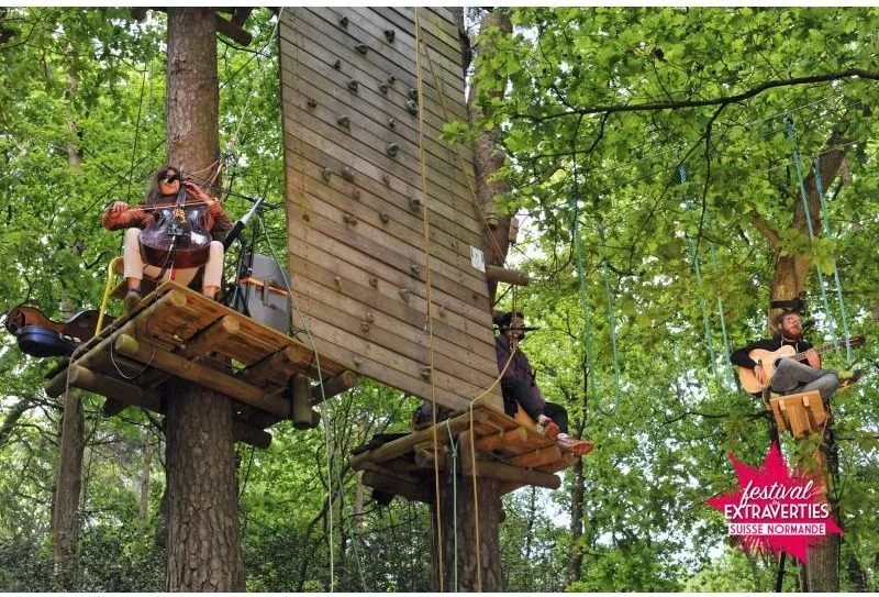 Trois personnes jouent de la guitare et chantent dans des plateformes suspendues dans les arbres lors d'un festival en Normandie.