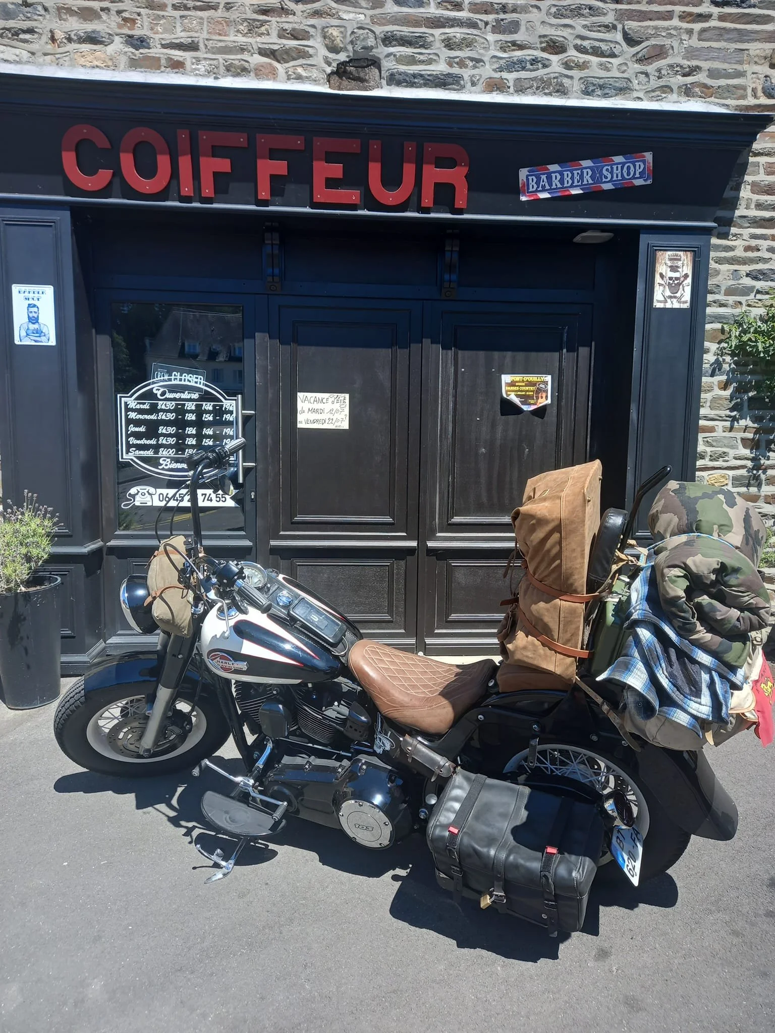Une moto stationnée devant un salon de coiffure nommé 'Coiffeur' à l'extérieur d'un bâtiment en pierre. La moto est équipée de sacs de voyage et de vêtements attachés à l'arrière.