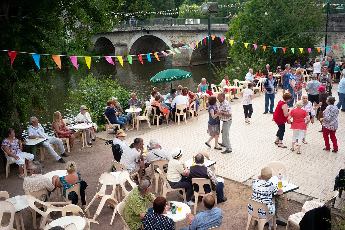 Fête Guinguette en plein air à Pont d'Ouilly en plein air près d'une rivière avec des personnes dansant, discutant et assises à des tables tandis que des drapeaux colorés décorent l'espace