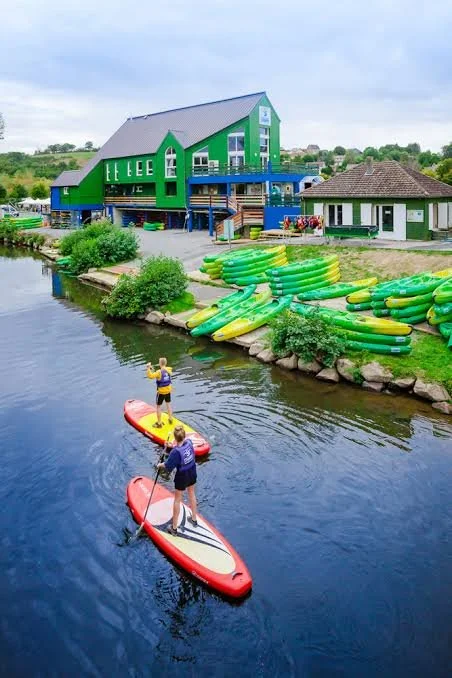 Deux personnes font du paddle sur une rivière près d'une zone avec plusieurs radeaux gonflables verts et jaunes empilés, avec un bâtiment vert et bleu en arrière-plan.