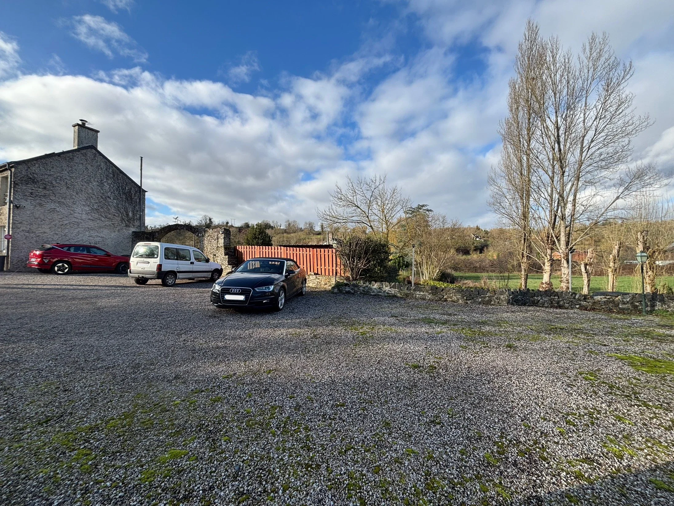 Un parking en plein air avec plusieurs voitures, un bâtiment ancien en pierre à gauche, un ciel partiellement nuageux, des arbres dénudés et un paysage rural en arrière-plan.